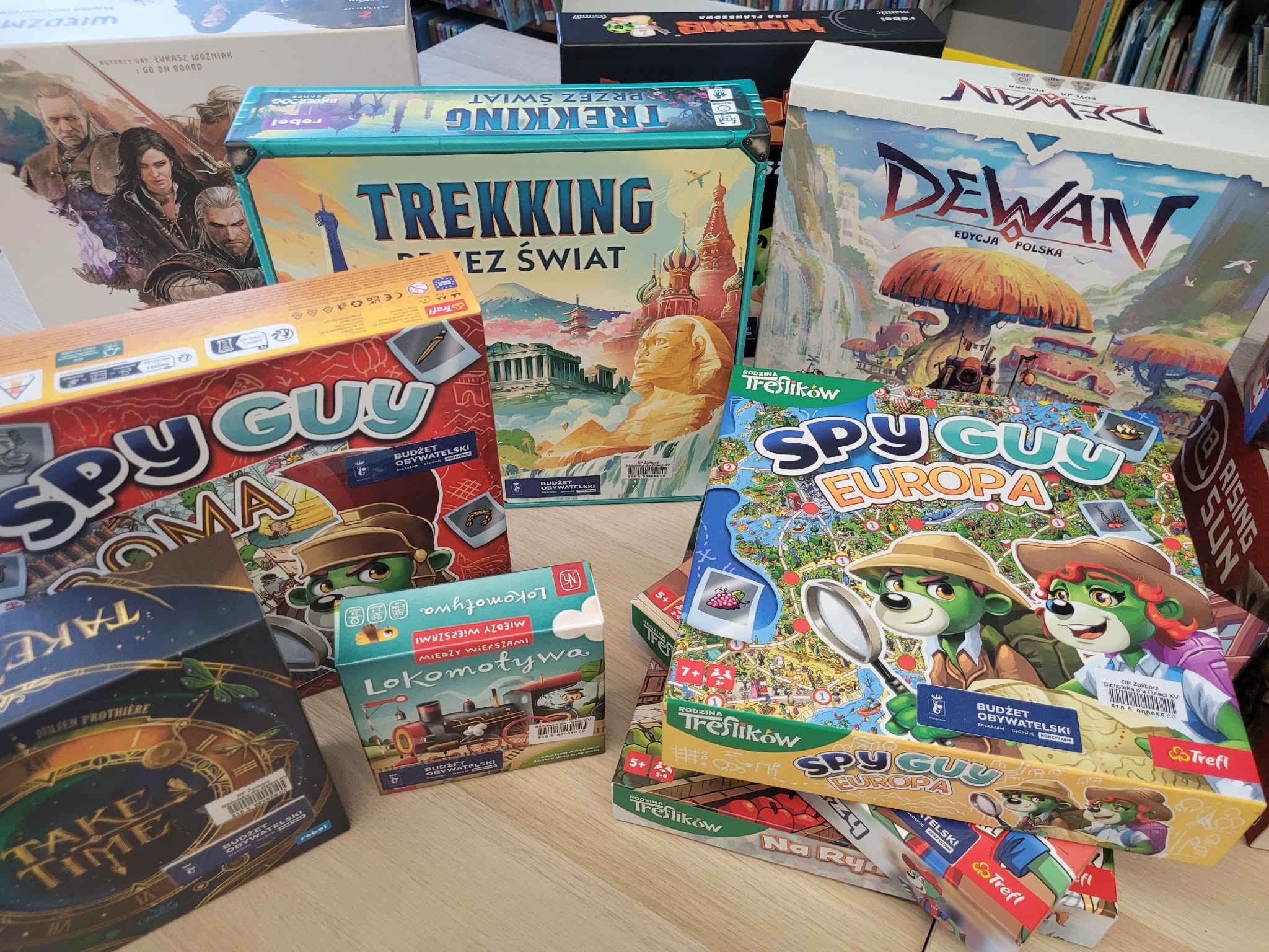 Na stole w bibliotece znajdują się gry planszowe z oznaczeniem „Budżet Obywatelski”, m.in. „Trekking przez świat”, „Dewan”, „Spy Guy Europa”, „Lokomotywa”, „Take a Thief” oraz „Rising Sun”. W tle widoczne półki z książkami.