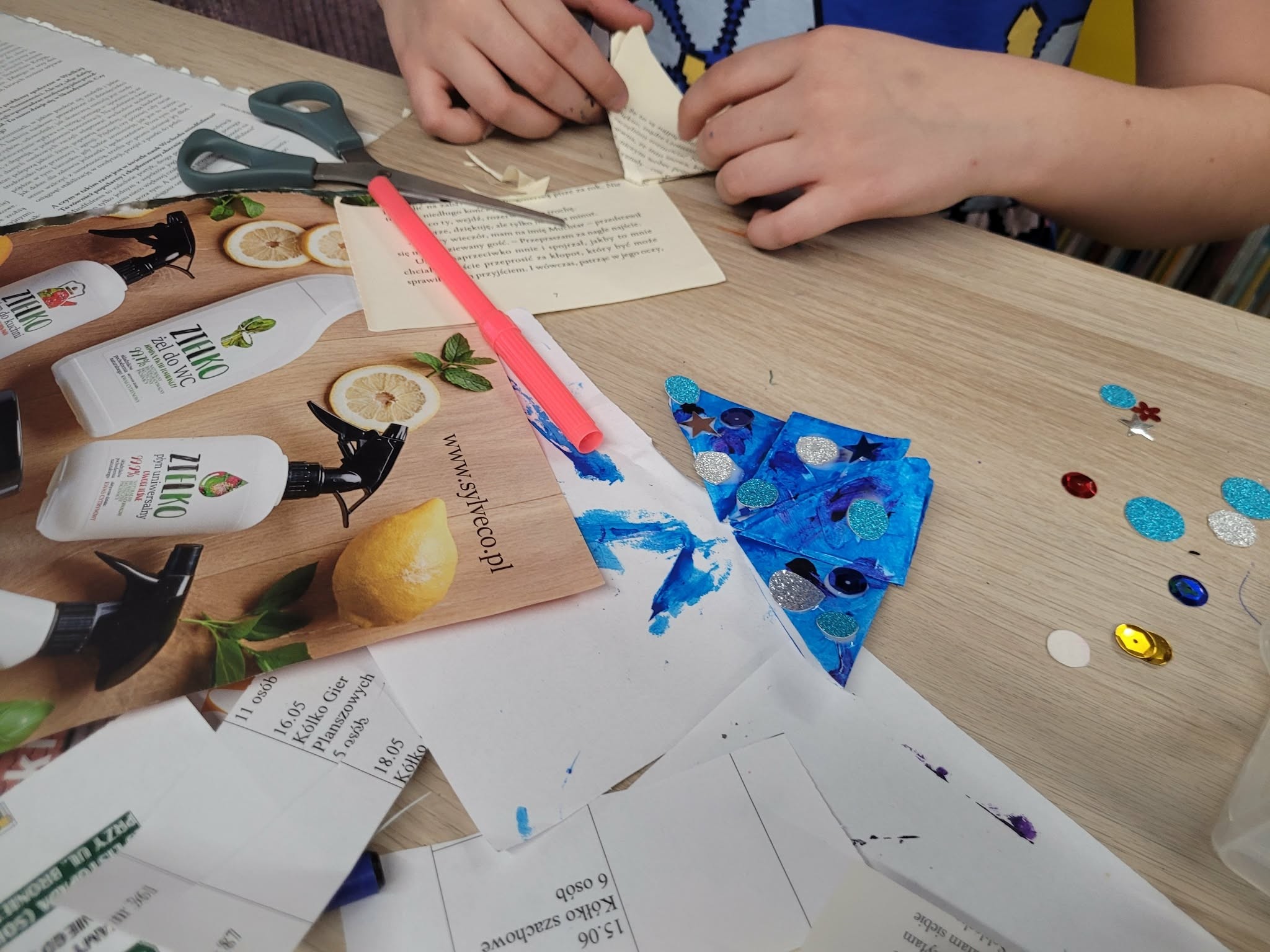 Dziecko składa papierowy kształt przy stole; obok widoczne nożyczki, kolorowe elementy dekoracyjne i niebieska papierowa praca plastyczna.