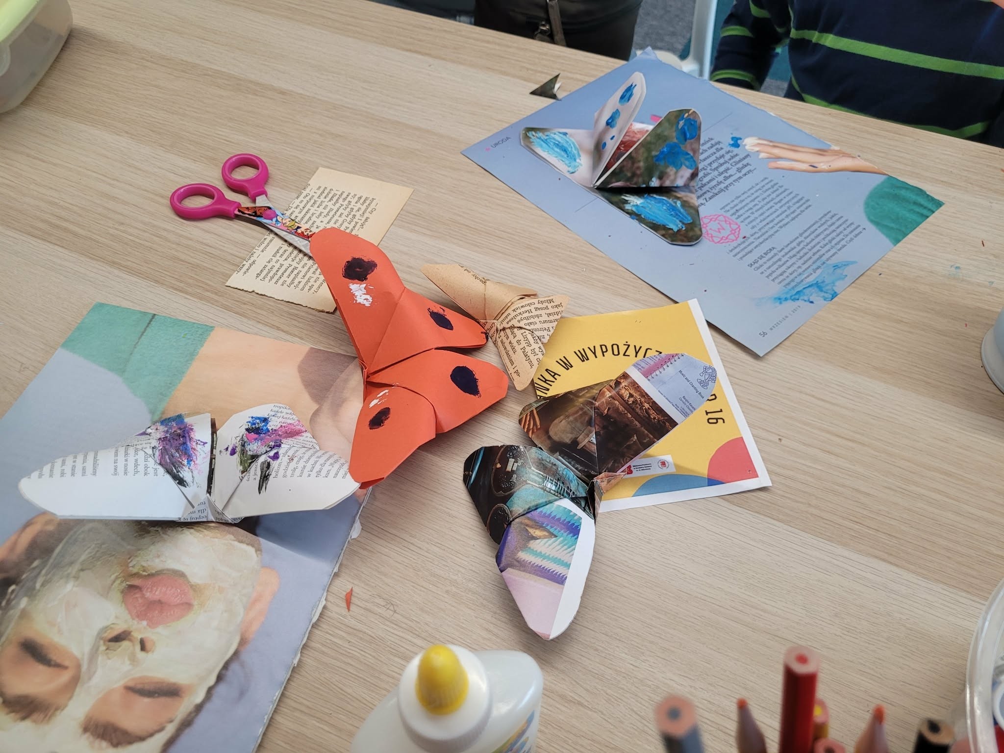Gotowe papierowe motyle ułożone na stole w bibliotece; obok nożyczki, klej i wycinki z czasopism.