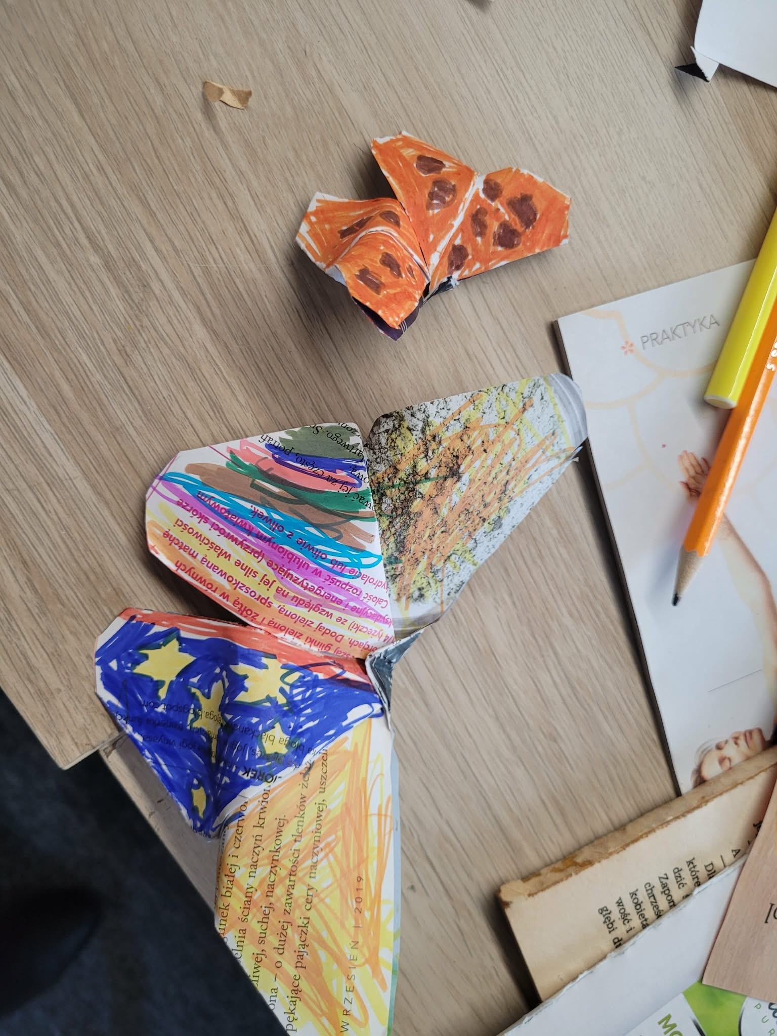 Papierowe motyle wykonane przez dzieci, ułożone na drewnianym stole; prace są kolorowane flamastrami i złożone z wyciętych fragmentów papieru.