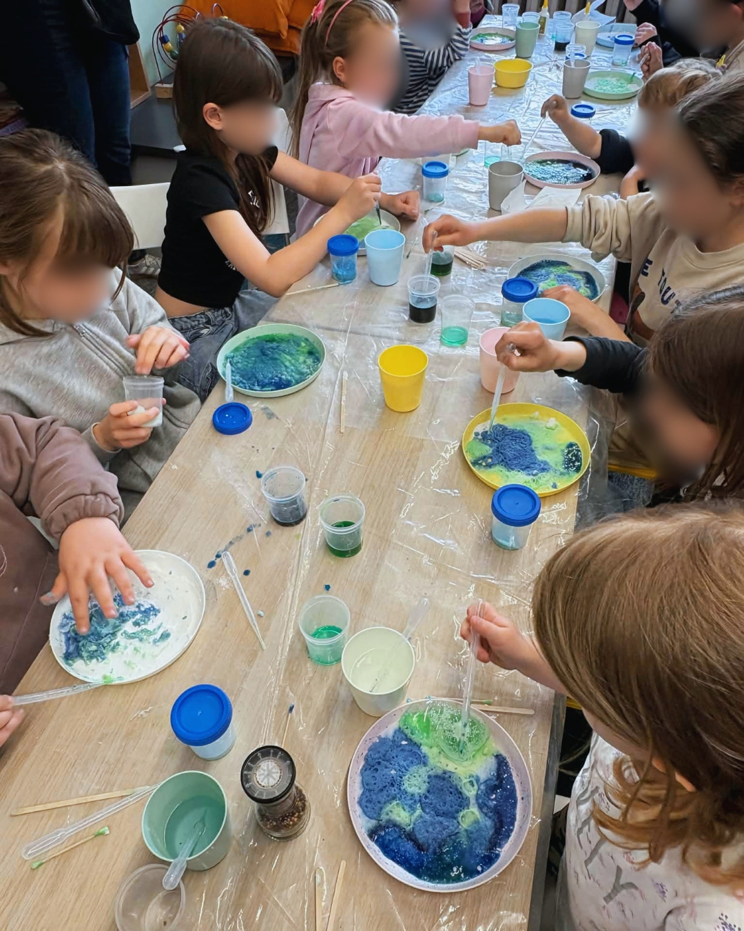 Grupa dzieci siedzi przy stole przykrytym folią i uczestniczy w zajęciach plastyczno-eksperymentalnych. Pod opieką dorosłej osoby mieszają w kubkach kolorowe substancje za pomocą pipet i patyczków. Na stole znajdują się małe pojemniki z barwnikami, klej i akcesoria do pracy.