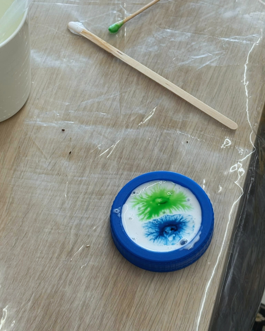 Na stole materiały do eksperymentów naukowych dla dzieci: kubeczek, drewniane patyczki, niebieska miseczka z substancją w kolorach białym, zielonym i niebieskim.