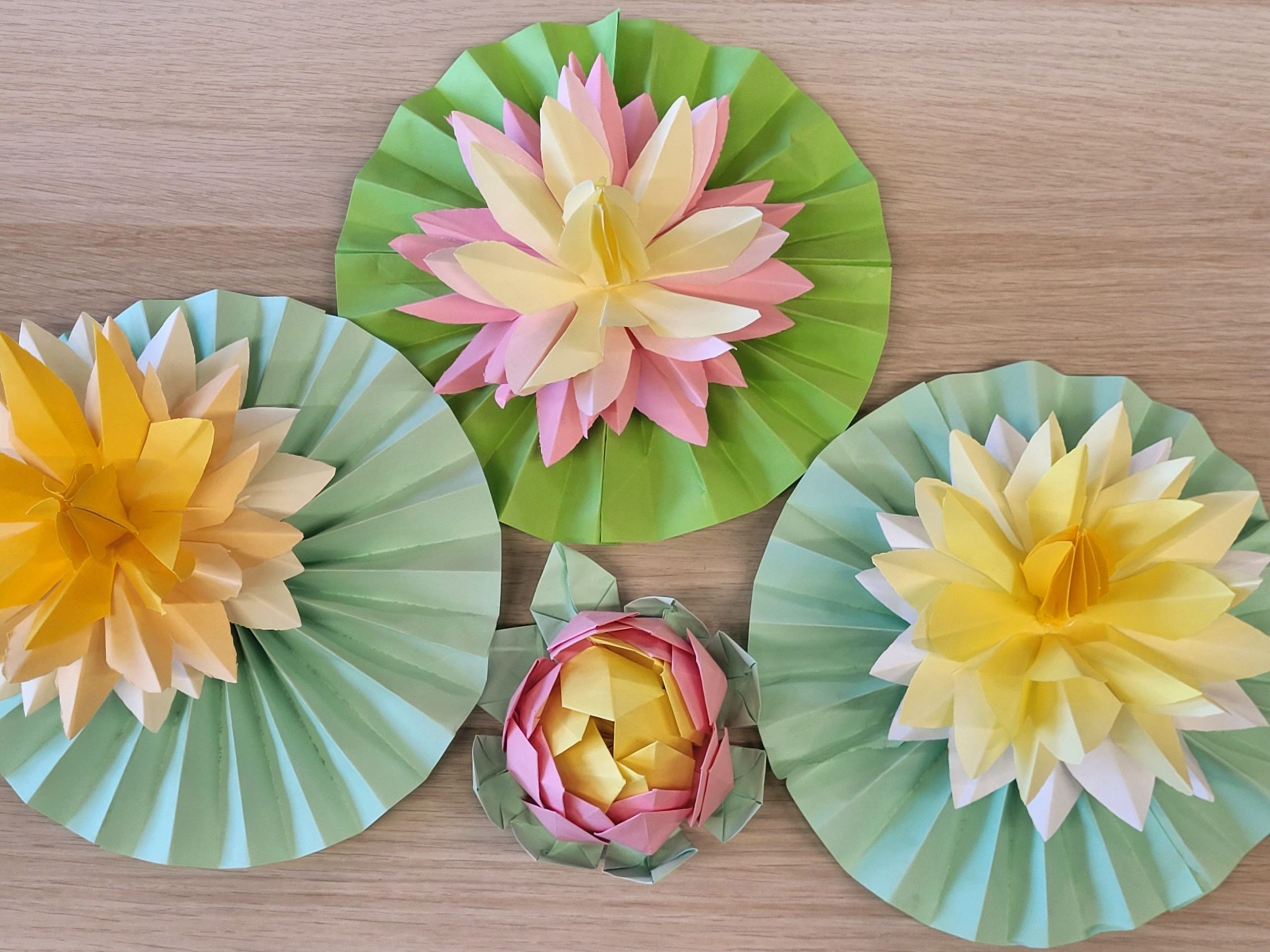 Kolorowe papierowe kwiaty origami przypominające lilie wodne. Trzy duże, rozkwitnięte kwiaty w odcieniach żółci, różu i kremu znajdują się na zielonych, harmonijkowo złożonych liściach. Pomiędzy nimi umieszczony jest mniejszy, zamknięty pąk w kolorach różowym i żółtym z zielonymi elementami. Całość ułożona na jasnym drewnianym tle.
