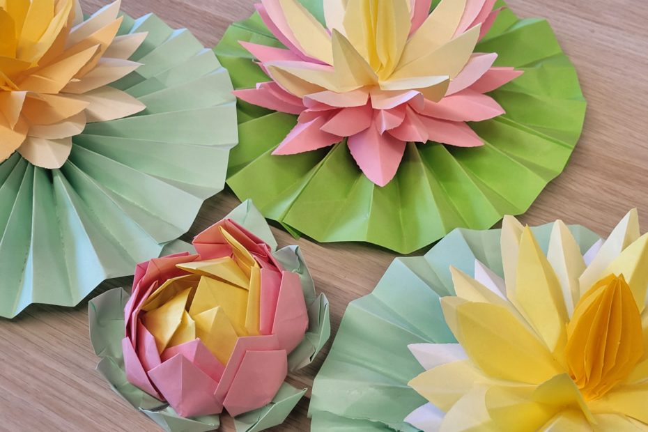 Zbliżenie na papierowe kwiaty origami w kształcie lilii wodnych. Widoczne są dwa duże kwiaty w odcieniach żółci i różu na zielonych, złożonych liściach oraz mniejszy, zamknięty pąk w kolorze różowo-żółtym. Kompozycja leży na jasnej, drewnianej powierzchni.