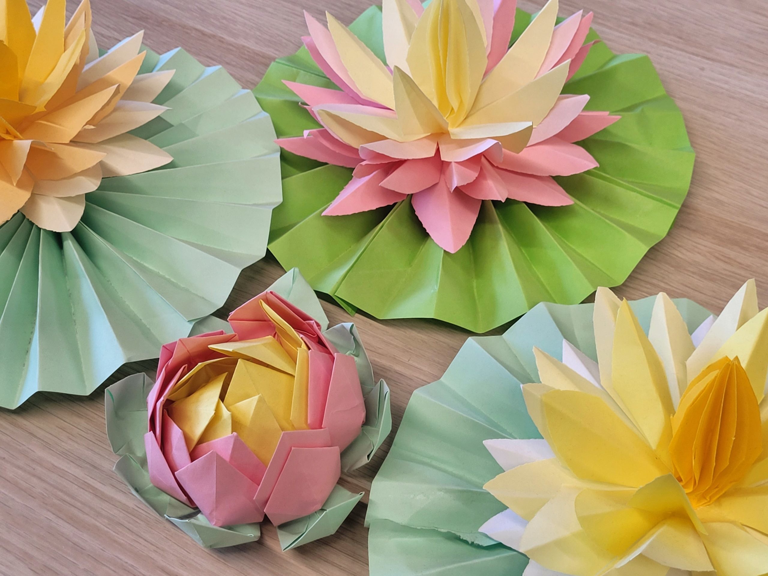 Zbliżenie na papierowe kwiaty origami w kształcie lilii wodnych. Widoczne są dwa duże kwiaty w odcieniach żółci i różu na zielonych, złożonych liściach oraz mniejszy, zamknięty pąk w kolorze różowo-żółtym. Kompozycja leży na jasnej, drewnianej powierzchni.
