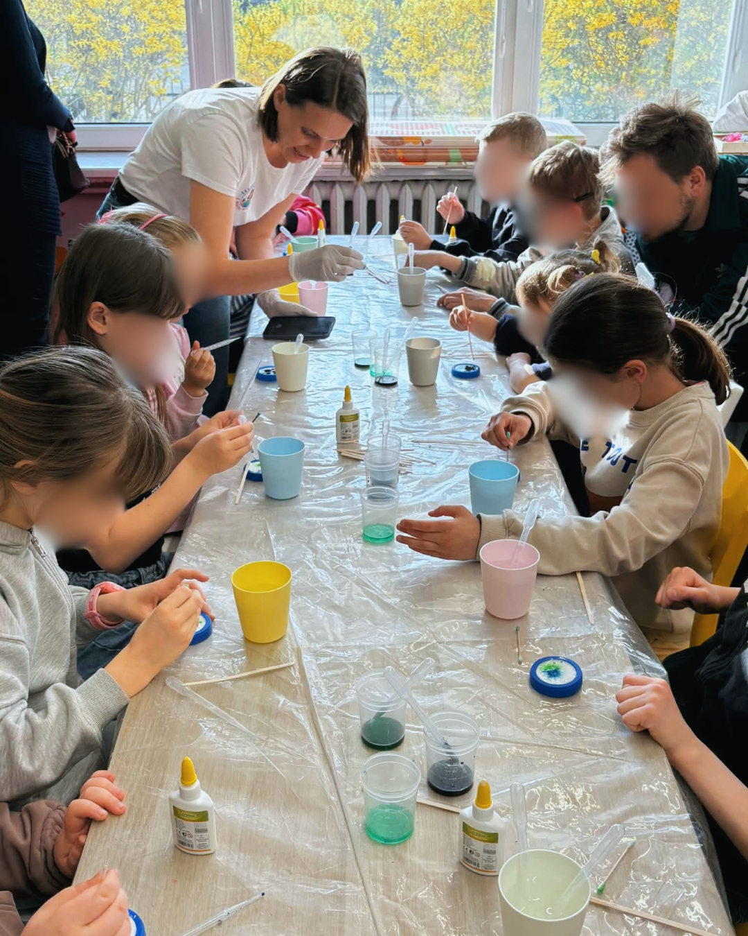 Grupa dzieci siedzi przy stole przykrytym folią i uczestniczy w zajęciach plastyczno-eksperymentalnych. Pod opieką dorosłej osoby mieszają w kubkach kolorowe substancje za pomocą pipet i patyczków. Na stole znajdują się małe pojemniki z barwnikami, klej i akcesoria do pracy. W tle widać duże okno z widokiem na drzewa.