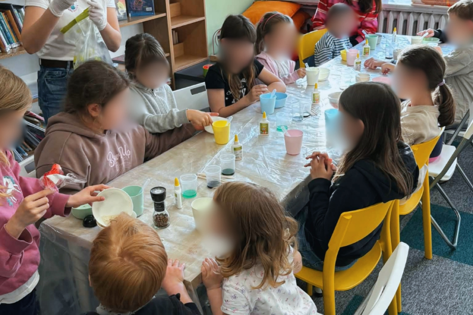 Grupa dzieci siedzi przy stole przykrytym folią i uczestniczy w zajęciach plastyczno-eksperymentalnych. Pod opieką dorosłej osoby mieszają w kubkach kolorowe substancje za pomocą pipet i patyczków. Na stole znajdują się małe pojemniki z barwnikami, klej i akcesoria do pracy. W tle widać duże okno z widokiem na drzewa.