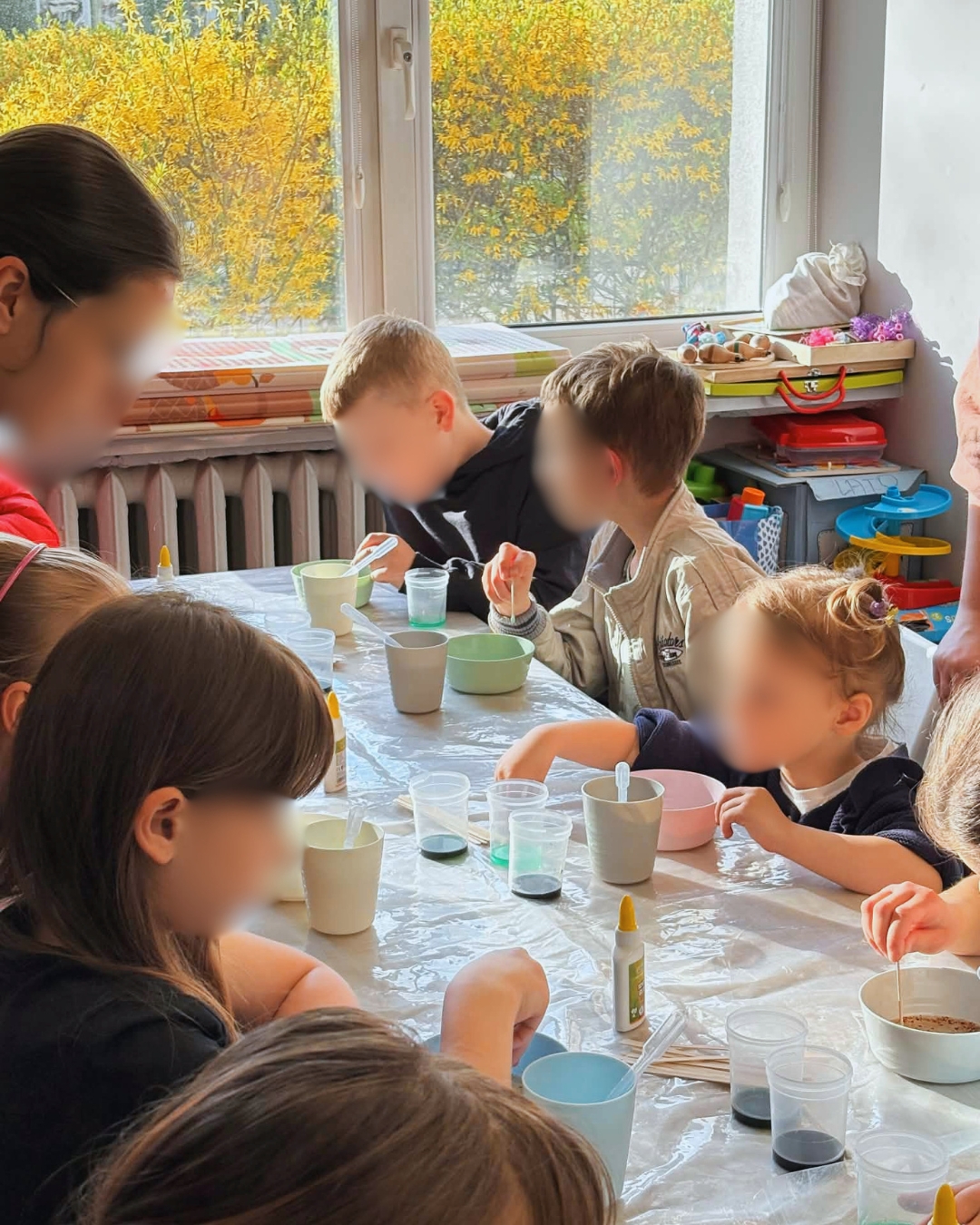 Grupa dzieci siedzi przy stole przykrytym folią i uczestniczy w zajęciach plastyczno-eksperymentalnych. Pod opieką dorosłej osoby mieszają w kubkach kolorowe substancje za pomocą pipet i patyczków. Na stole znajdują się małe pojemniki z barwnikami, klej i akcesoria do pracy. W tle widać duże okno z widokiem na drzewa.