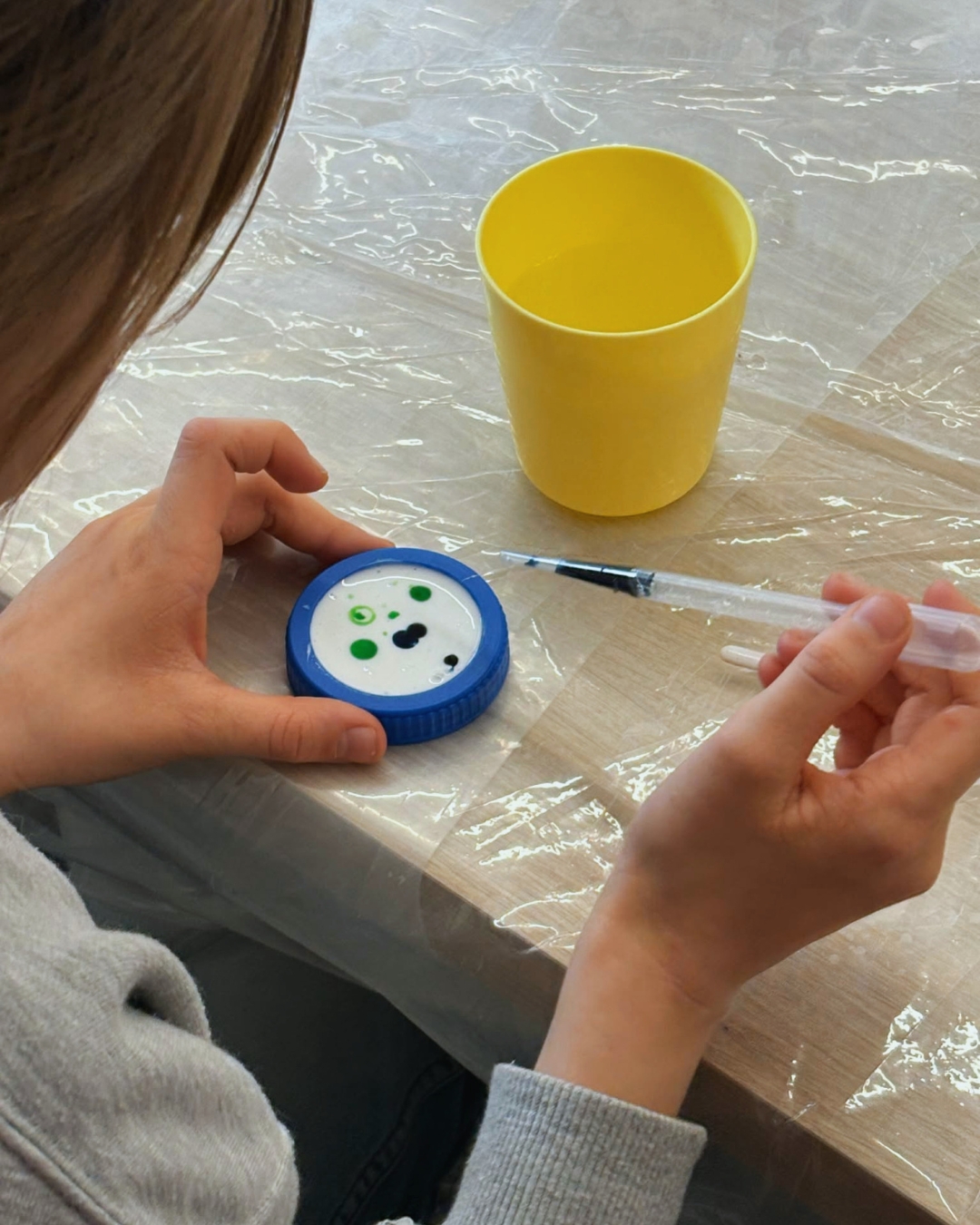 Dziecko siedzi przy stole zabezpieczonym folią i za pomocą pipety nakrapia kolorowe krople na białą powierzchnię w plastikowej nakrętce. Obok stoi żółty kubek z wodą.