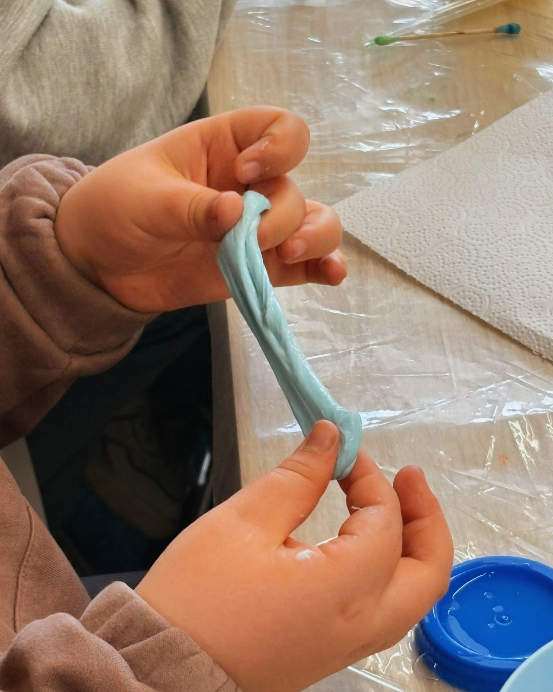 Dziecko trzyma w rękach niebieski slime. Siedzi przy stole pokrytym przezroczystą folią, na stole leży papierowa serwetka.