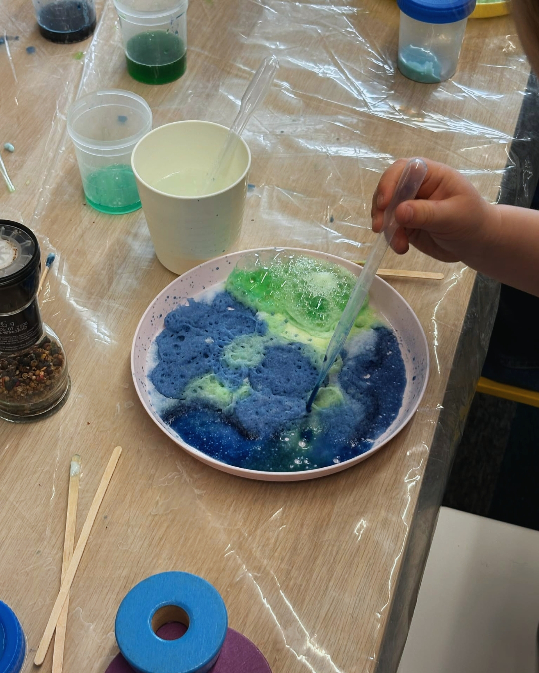 Dziecko trzyma pipetę i miesza pieniącą się, kolorową substancję w płaskim naczyniu. Na stole wokół znajdują się kubki, patyczki i pojemniki z barwnikami.
