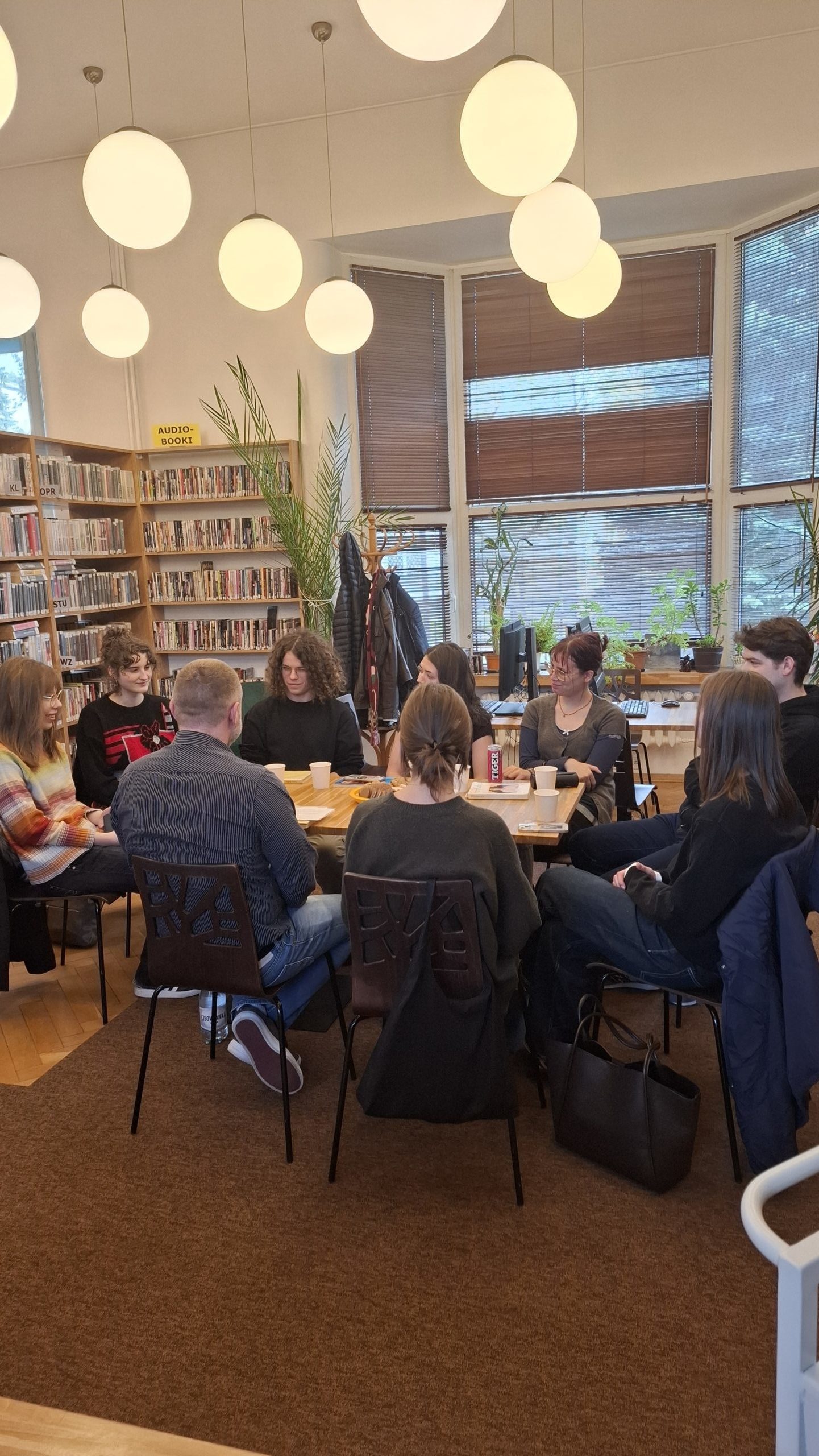 Grafika wyróżniająca przedstawiająca uczestników Młodzieżowego Dyskusyjnego Klubu Książki. Klubowicze siedzą wokół drewnianego stołu, omawiają książkę "Proces" F Kafki