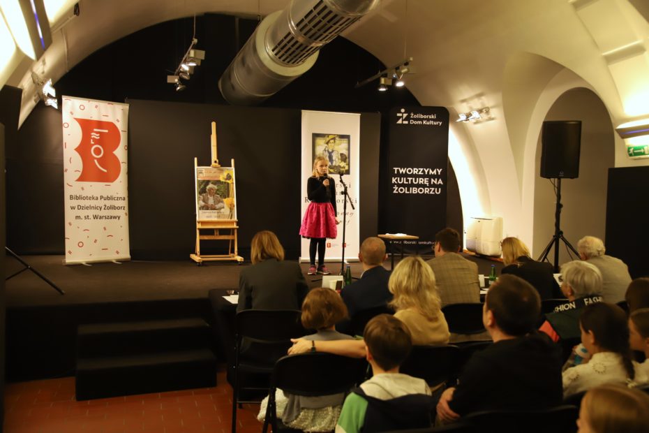 Zdjęcie uczestniczki Żoliborskiego konkursu recytatorskiego. Na scenie dziewczynka z mikrofonem, w tle baner Żoliborski Konkurs Recytatorski, Żoliborski Dom Kultury - Tworzymy Kulturę na Żolibori baner Biblioteka Publiczna w Dzielnicy Żoliborz, czarne tło. W sali siedzą słuchacze, jurorzy i uczestnicy odwróceni plecami.