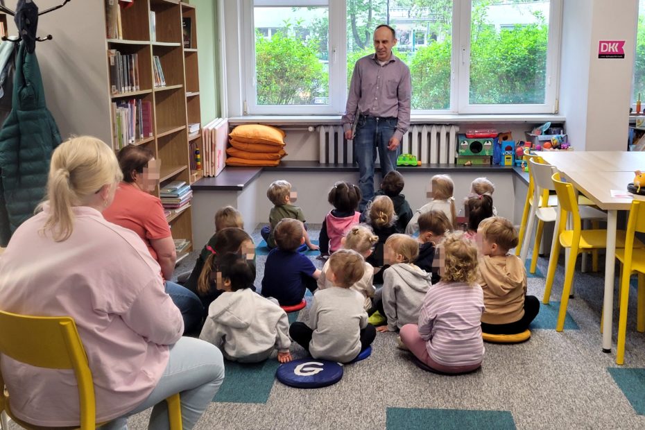 Grupa dzieci na spotkaniu w bibliotece, siedzą na poduszkach na dywanie, obok na krześle siedzi opiekunka. Przy oknie stoi bibliotekarz.