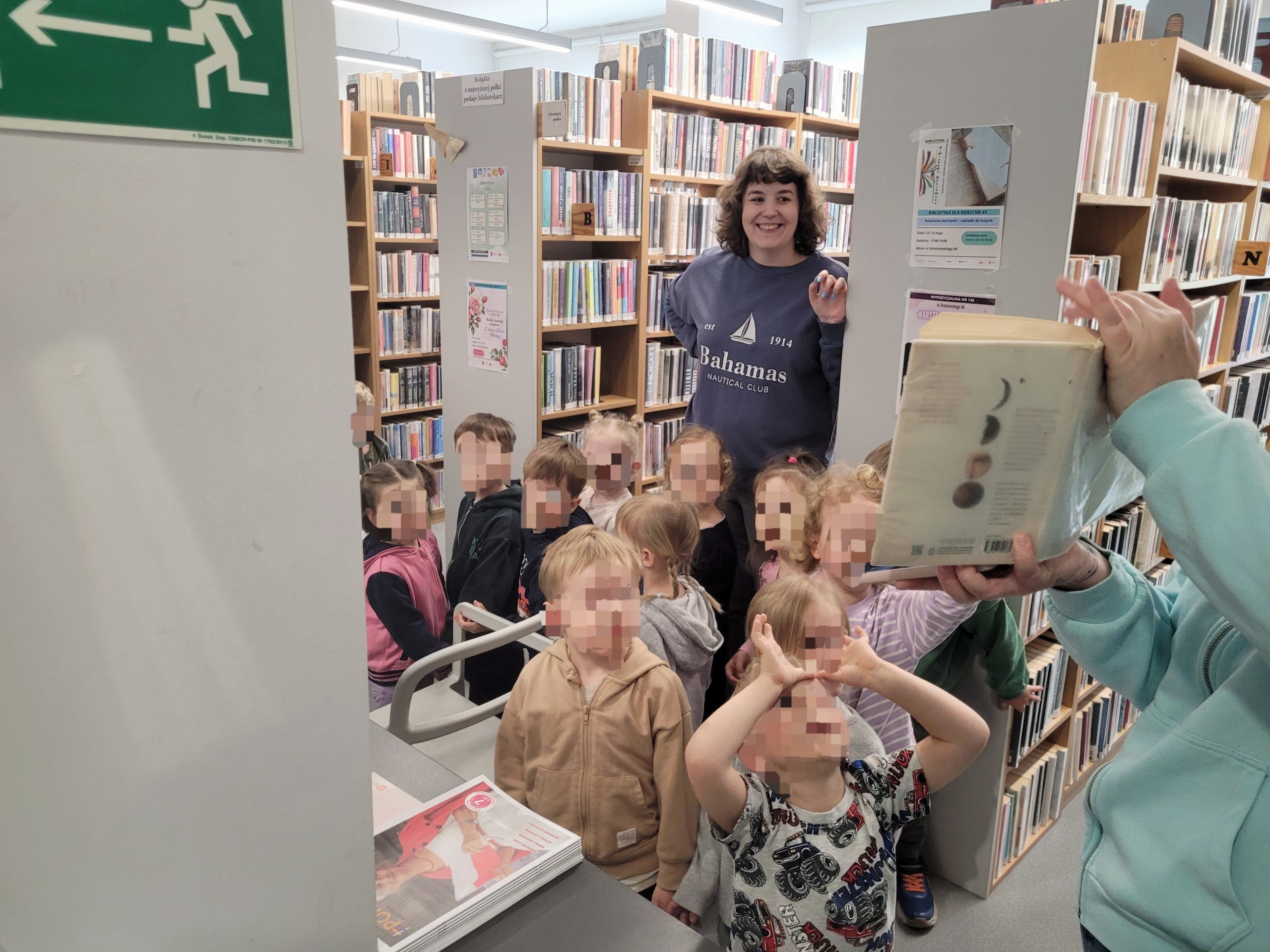 Grupa dzieci z kilkoma opiekunkami na spotkaniu w bibliotece stoją przy regałach z ksiązkami,