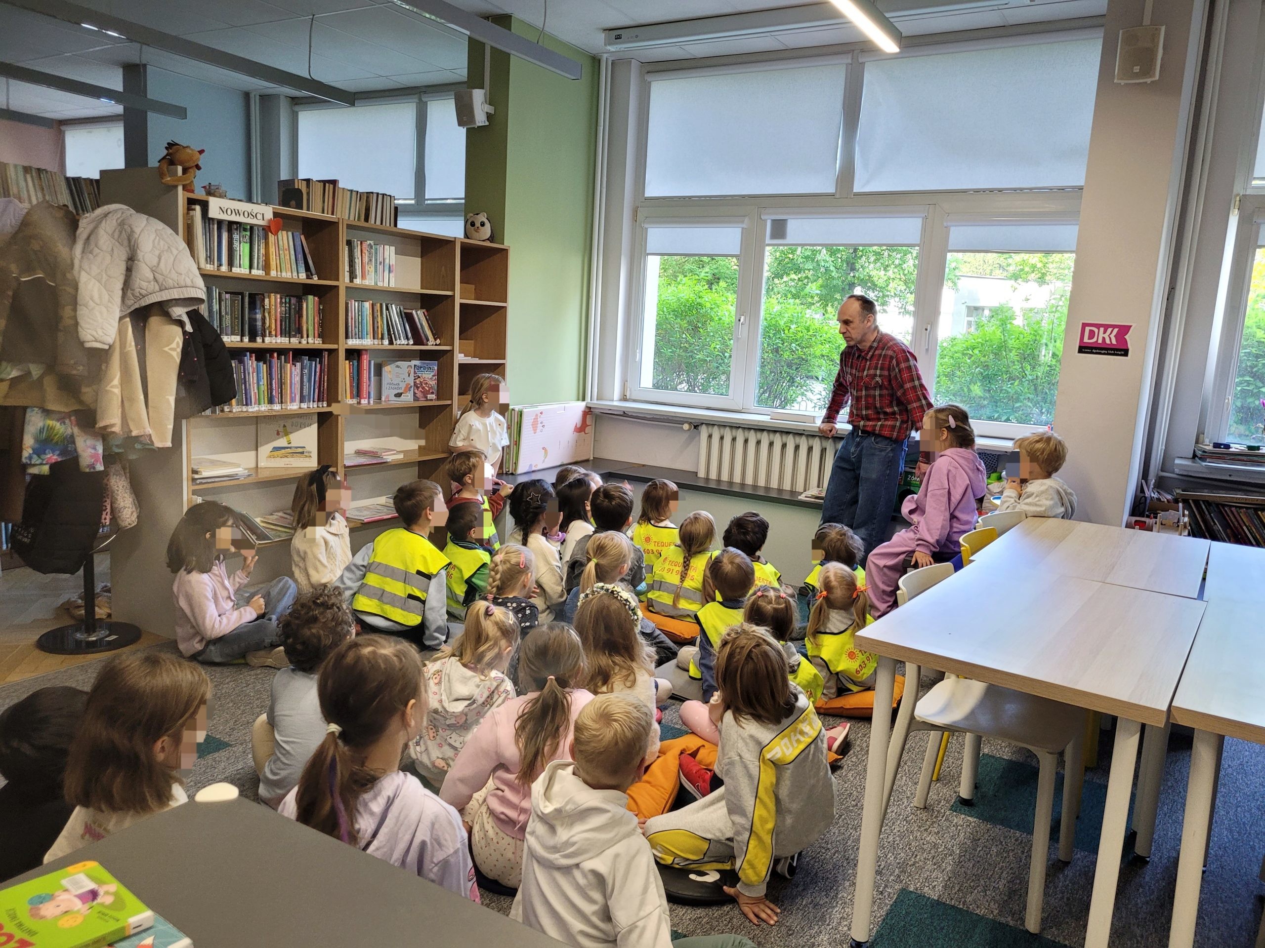 Grupa dzieci na spotkaniu w bibliotece, siedzą na poduszkach na dywanie. Przy oknie stoi bibliotekarz.