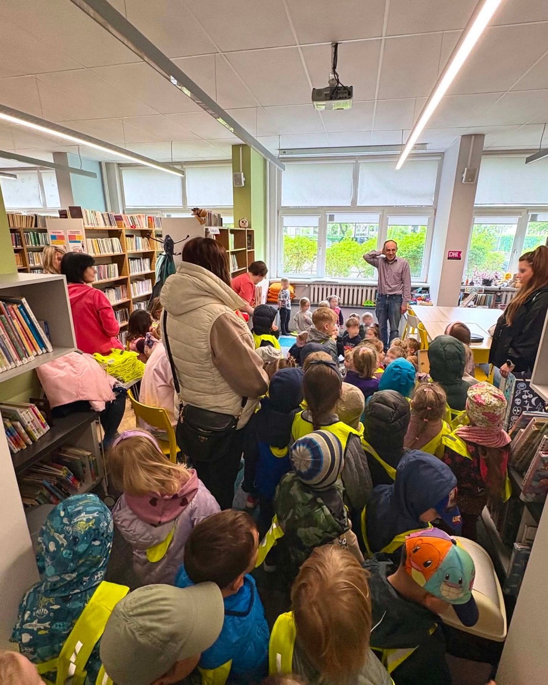 Grupa dzieci z kilkoma opiekunkami na spotkaniu w bibliotece. Przy oknie stoi bibliotekarz.
