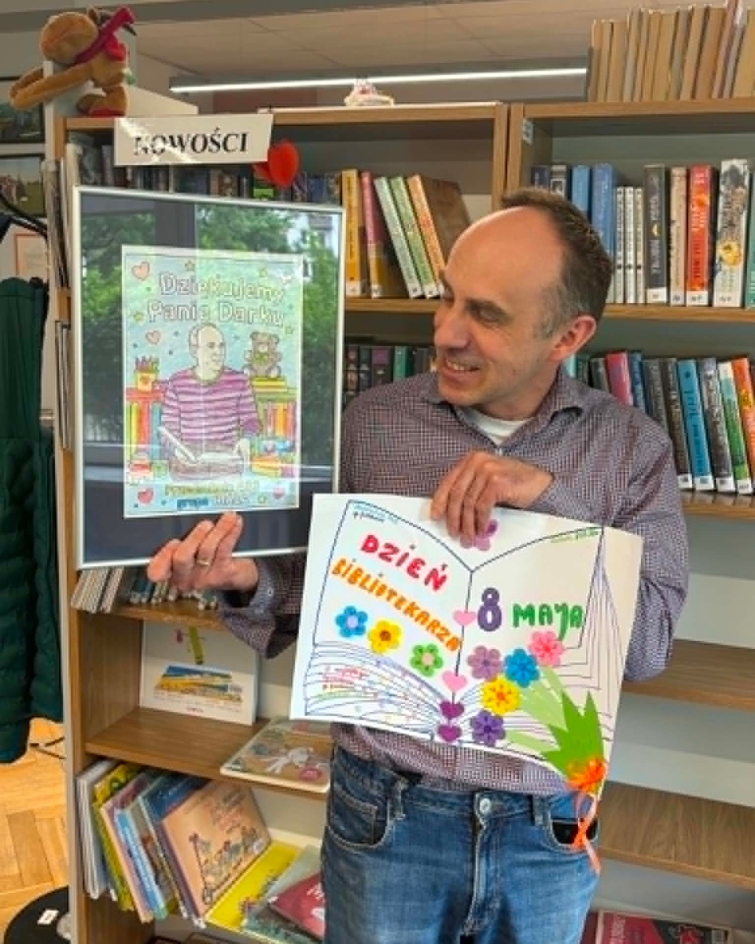 Bibliotekarz na tle regału z książkami trzyma w rękach portret zrobiony przez dzieci z napisem Dziękujemy, Panie Darku oraz wykonaną przez dzieci kartkę Dzień Bibliotekarza 8 maja.
