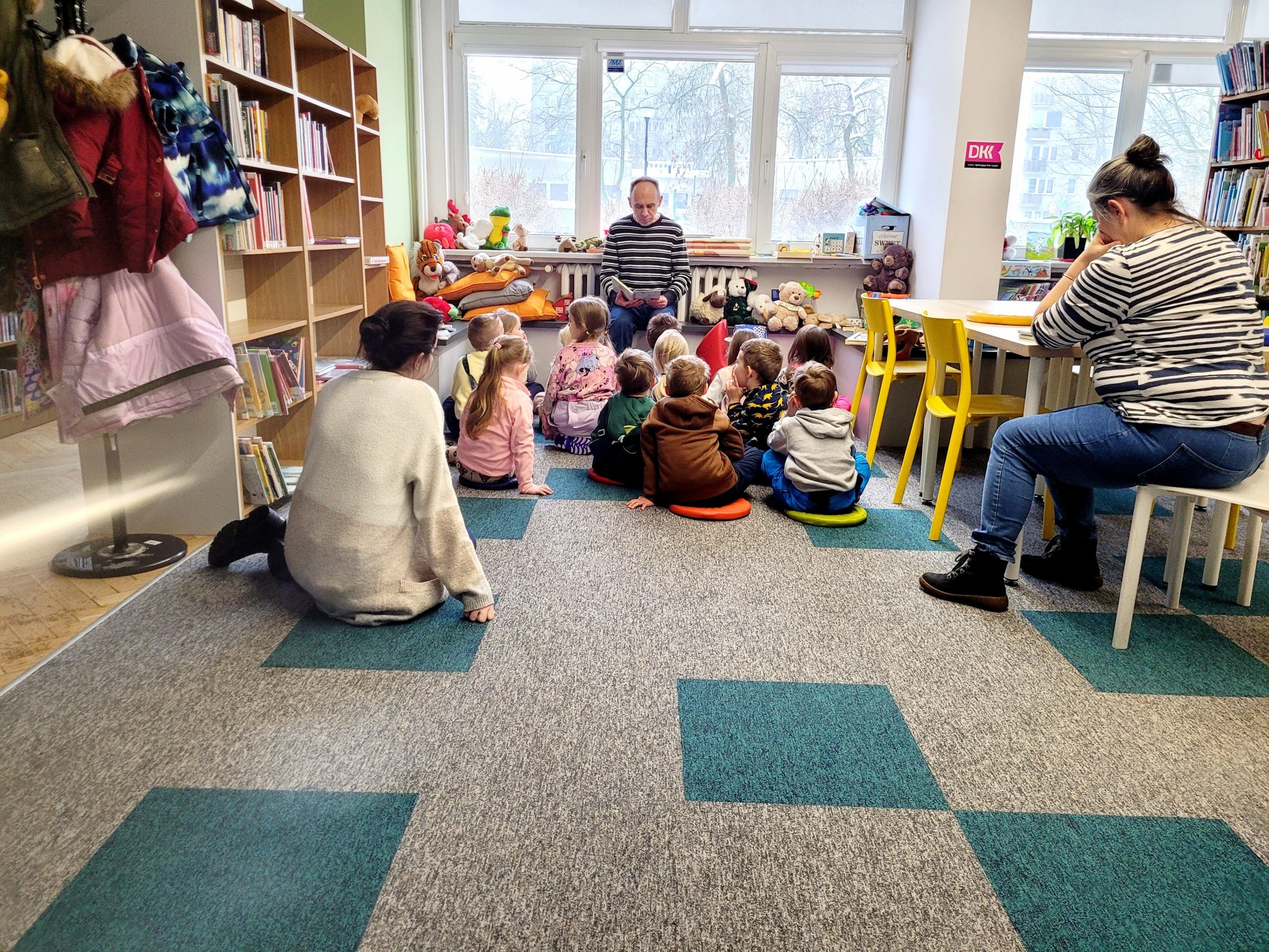 Grupa dzieci siedzi na dywanie w bibliotece i słucha mężczyzny, który czyta im książkę. Po lewej stronie na regale wiszą zimowe kurtki, a w tle za oknem widać zimowy krajobraz.