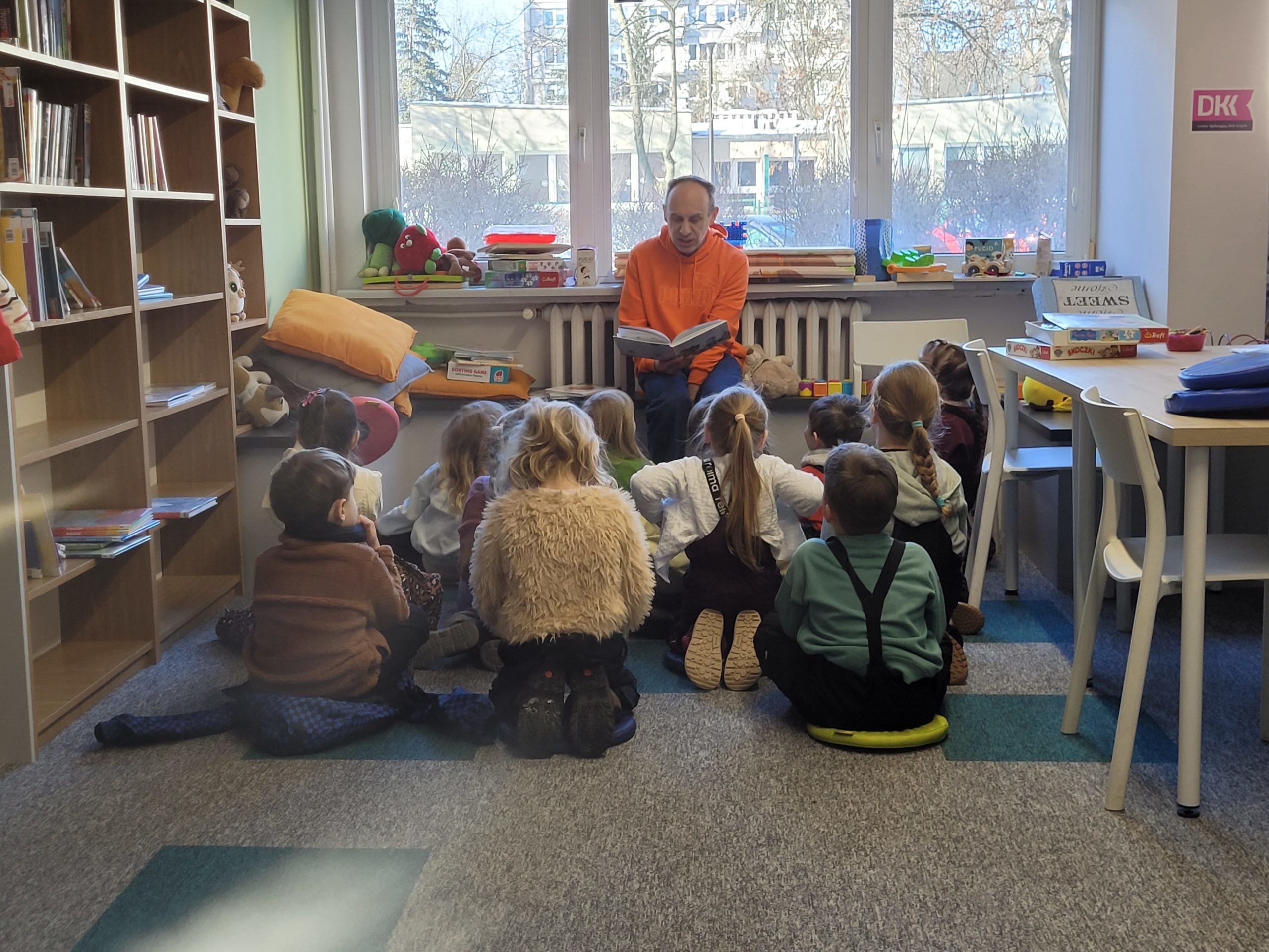 Mężczyzna w pomarańczowej bluzie z kapturem siedzi pod oknem w bibliotece i czyta książkę grupie dzieci siedzących przed nim na dywanie. Na parapecie leżą maskotki i gry planszowe.