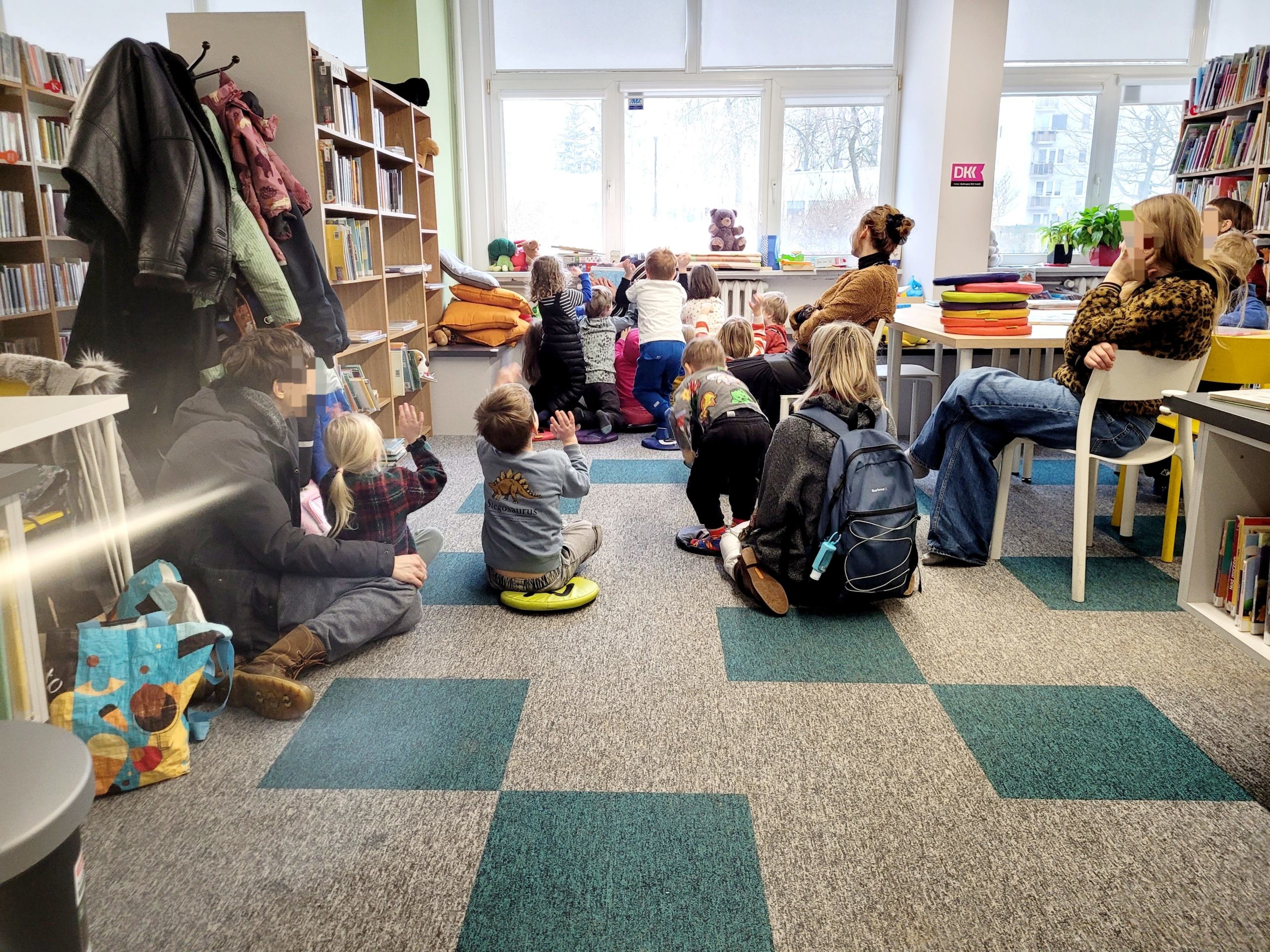 Spotkanie dla dzieci w bibliotece. Część dzieci i dorosłych siedzi na dywanie, a kilka osób stoi przy oknie podnosząc ręce. Po prawej stronie widoczne są stoliki i krzesła.