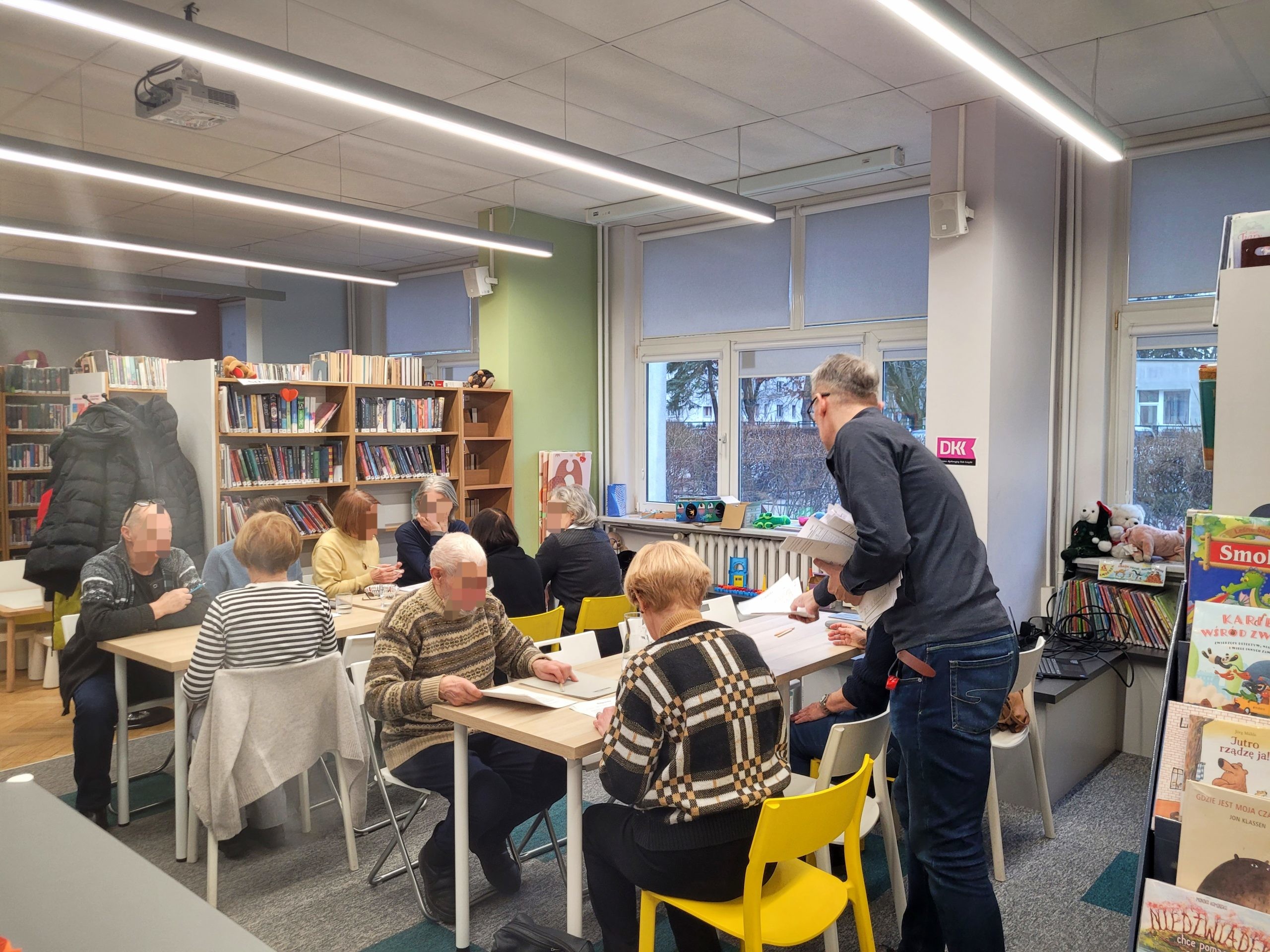 Grupa seniorów uczestniczy w warsztatach edukacyjnych w jasnym wnętrzu biblioteki. Uczestnicy siedzą przy połączonych jasnych stołach, na których leżą materiały szkoleniowe. Część osób przegląda dokumenty, inni rozmawiają. Między stołami stoi prowadzący – mężczyzna w okularach i ciemnym, pasiastym swetrze, trzymający w rękach notatki i rozmawiający z jedną z uczestniczek. W tle widoczne są regały pełne książek oraz duże okna.