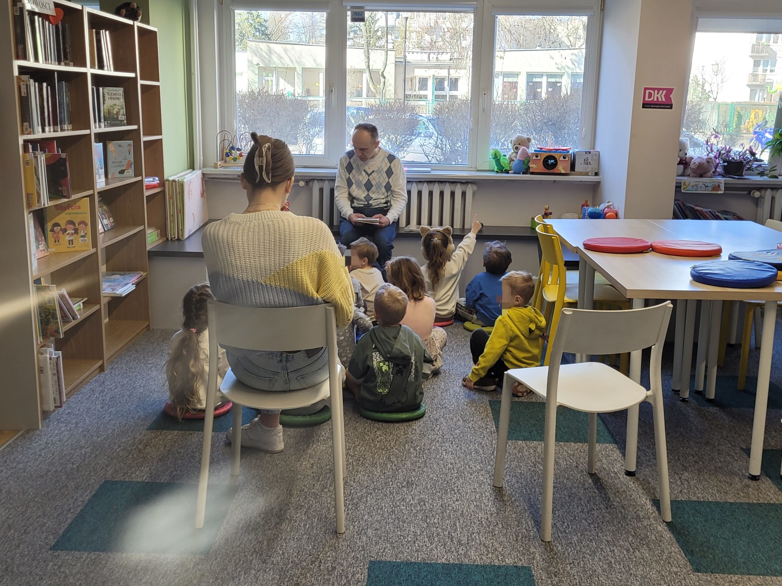 Mężczyzna w jasnym swetrze w romby czyta książkę dzieciom zgromadzonym w kąciku dla dzieci w bibliotece. Tyłem do obiektywu siedzi kobieta na krześle, towarzysząc grupie.