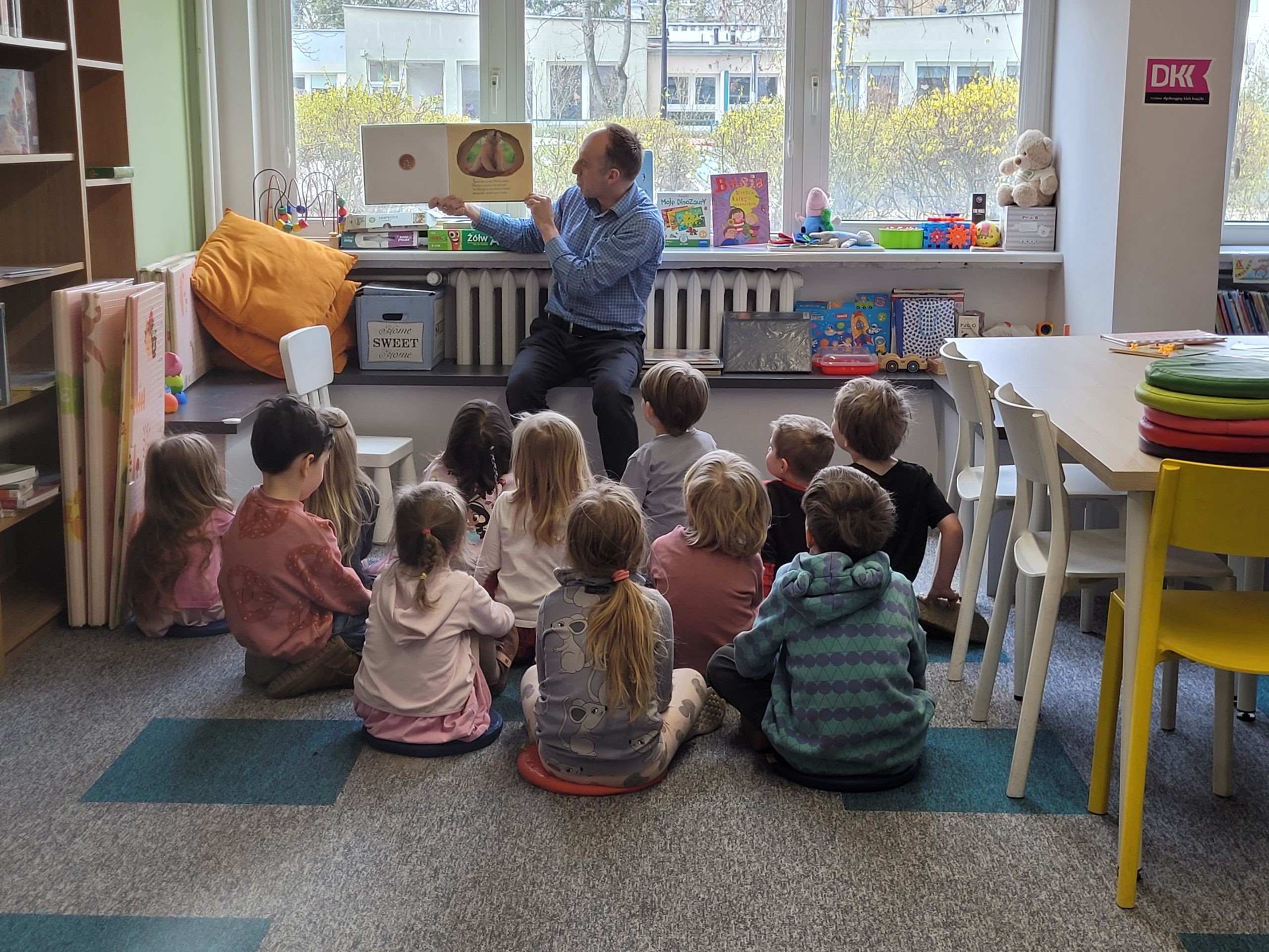 Prowadzący siedzi na niskim podeście pod oknem biblioteki i pokazuje dzieciom ilustrację w otwartej, dużej książce. Dzieci siedzą tyłem na kolorowych poduszkach i patrzą na obrazek.