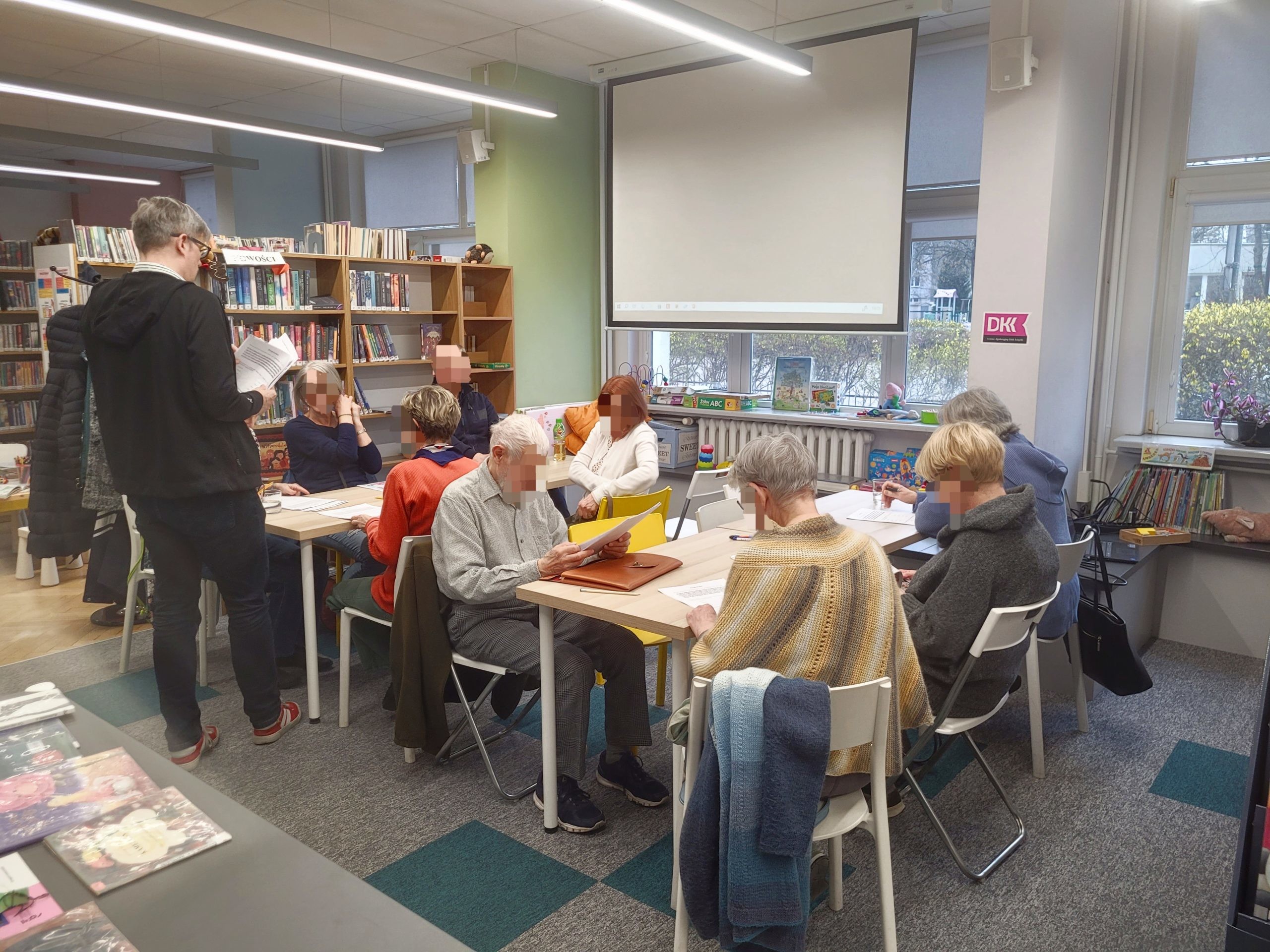 Spotkanie edukacyjne dla seniorów w bibliotece. Uczestnicy siedzą przy stołach ustawionych w rzędzie pod oknem, skupieni na czytaniu rozdanych materiałów lub wypełnianiu ćwiczeń. Po lewej stronie, tyłem do obiektywu, stoi prowadzący spotkanie w ciemnej bluzie i okularach, trzymający kartki papieru. Nad oknem zwinięty jest biały ekran projekcyjny. W tle widać regały z książkami.