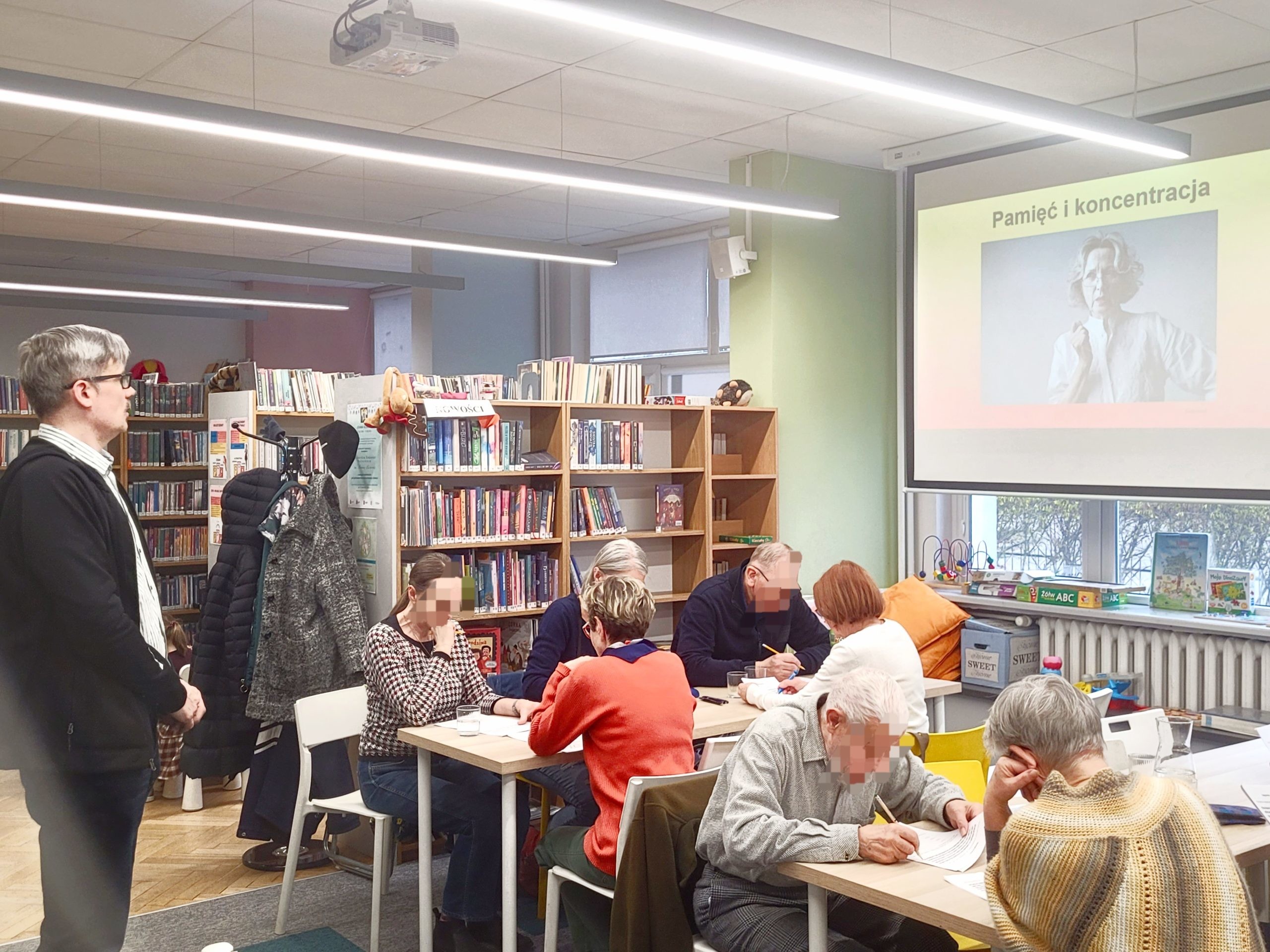 Warsztaty dla seniorów w bibliotece zatytułowane „Pamięć i koncentracja”. Na dużym ekranie projekcyjnym wyświetlany jest slajd z tym tytułem oraz czarno-białym portretem starszej kobiety. Przy stołach siedzą starsze osoby, które piszą w swoich arkuszach ćwiczeń lub dyskutują. Po lewej stronie stoi prowadzący w okularach i rozpiętej czarnej bluzie, obserwując pracę grupy.