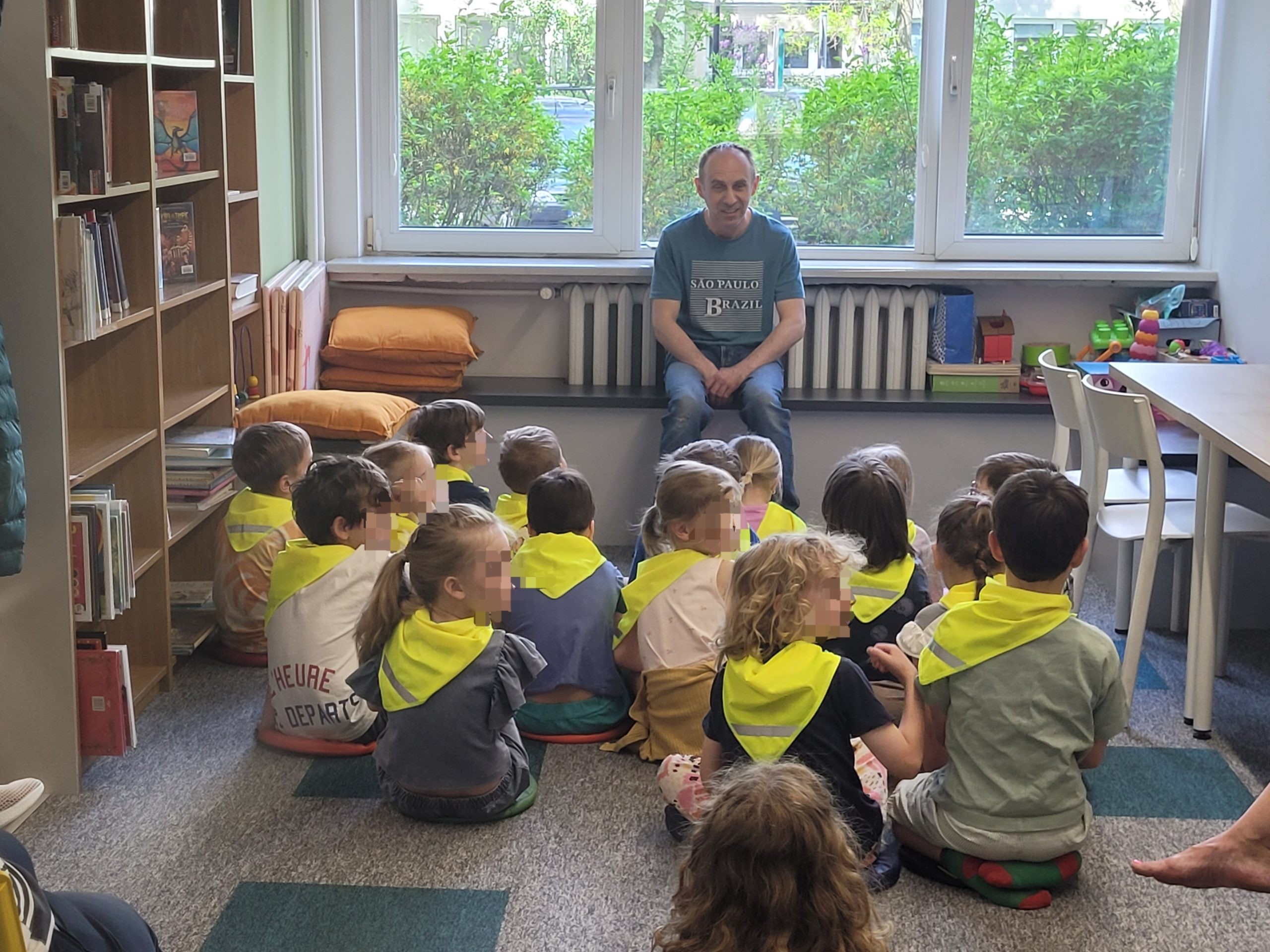 Mężczyzna w niebieskiej koszulce z napisem rozmawia z grupą dzieci siedzących na dywanie w bibliotece. Wszystkie dzieci mają na sobie jaskrawożółte kamizelki lub chusty odblaskowe. Za oknem widać zielone krzewy.