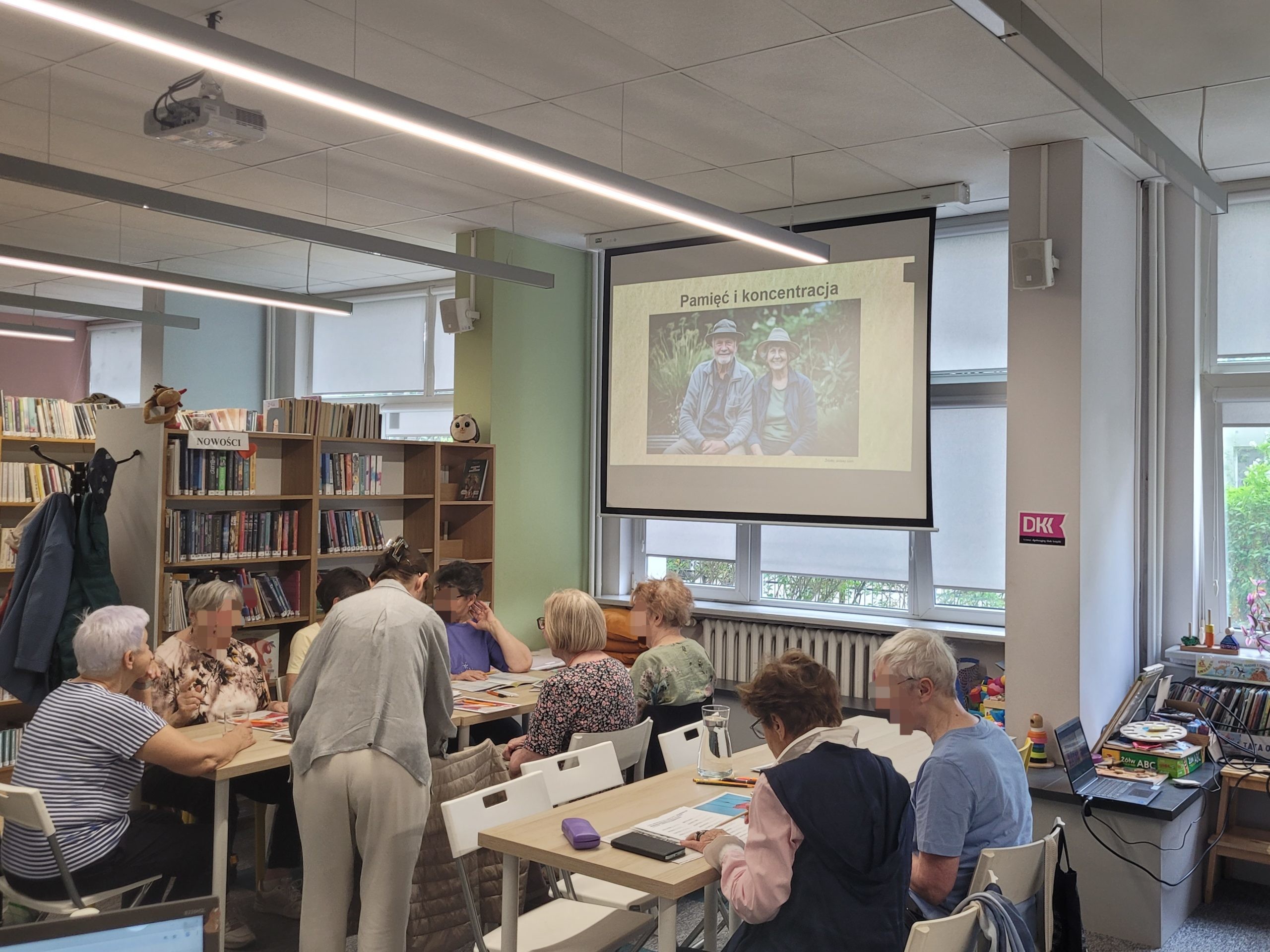 Seniorzy biorą udział w zajęciach przybliżających temat pamięci i koncentracji w bibliotece. Na ekranie w tle wyświetlany jest slajd z napisem „Pamięć i koncentracja” oraz zdjęciem uśmiechniętej pary starszych ludzi na łonie natury. Uczestnicy siedzą przy dwóch stołach – jedni piszą, inni rozmawiają z instruktorką w jasnym ubraniu, która pochyla się nad ich stolikiem.