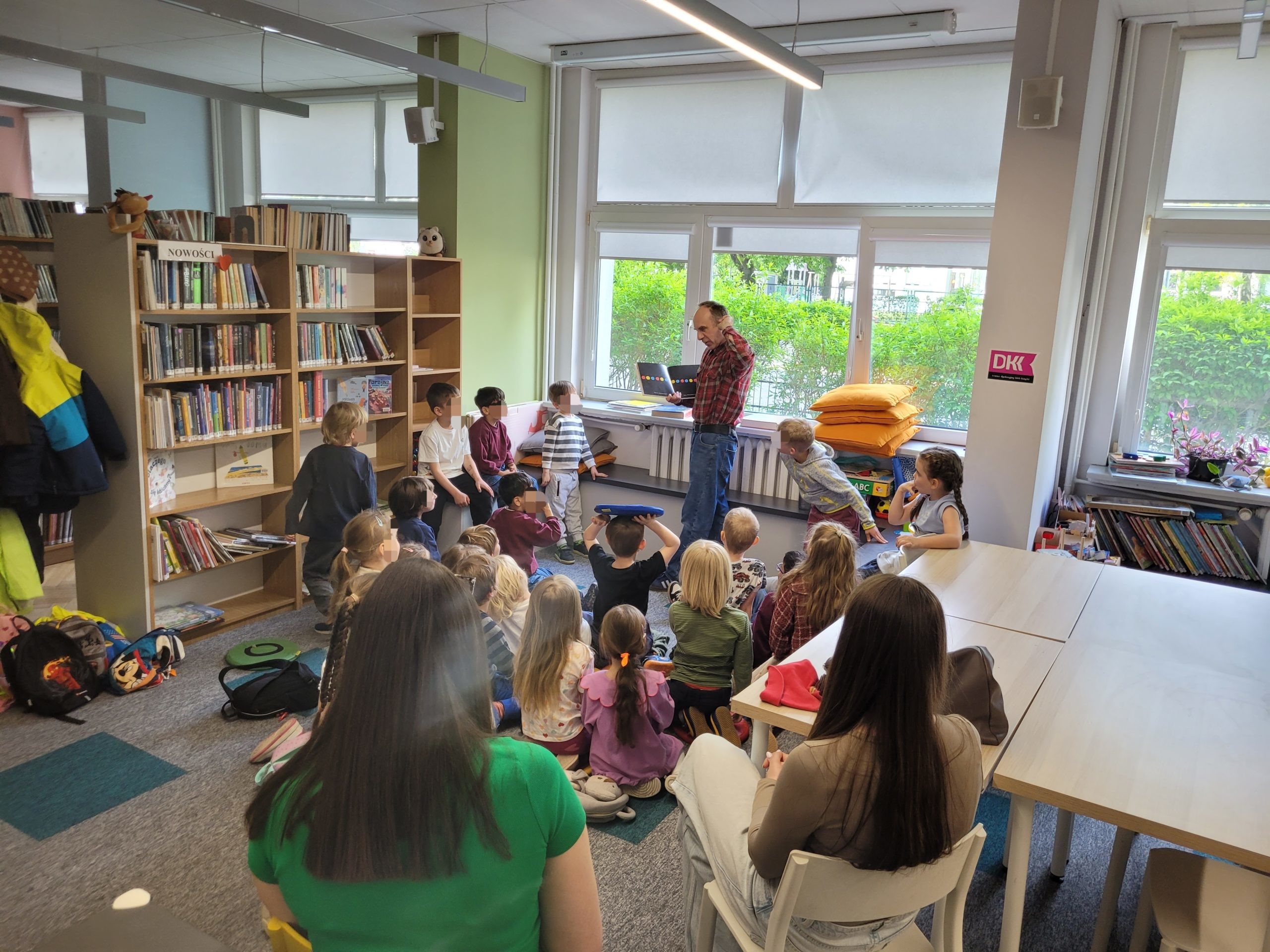 Spotkanie w jasnej sali bibliotecznej. Mężczyzna w koszuli w kratę stoi przy oknie i pokazuje dzieciom książkę z kolorowymi kropkami na stronach. Na pierwszym planie widoczne są dwie siedzące tyłem kobiety.