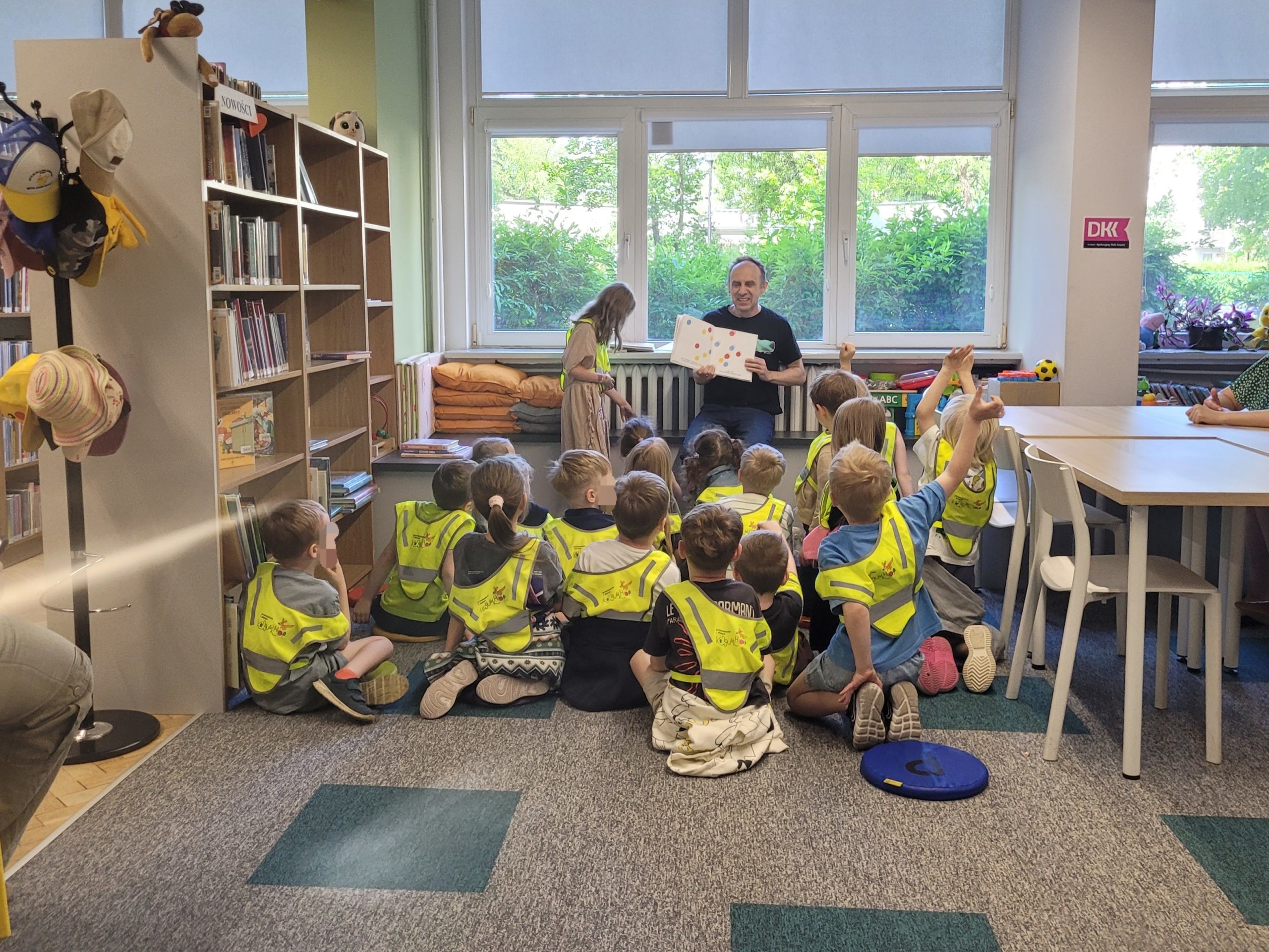 Mężczyzna pokazuje dzieciom otwartą książkę z kolorowymi punktami. Dzieci w żółtych kamizelkach odblaskowych siedzą na dywanie w bibliotece, jedno z nich podnosi rękę.