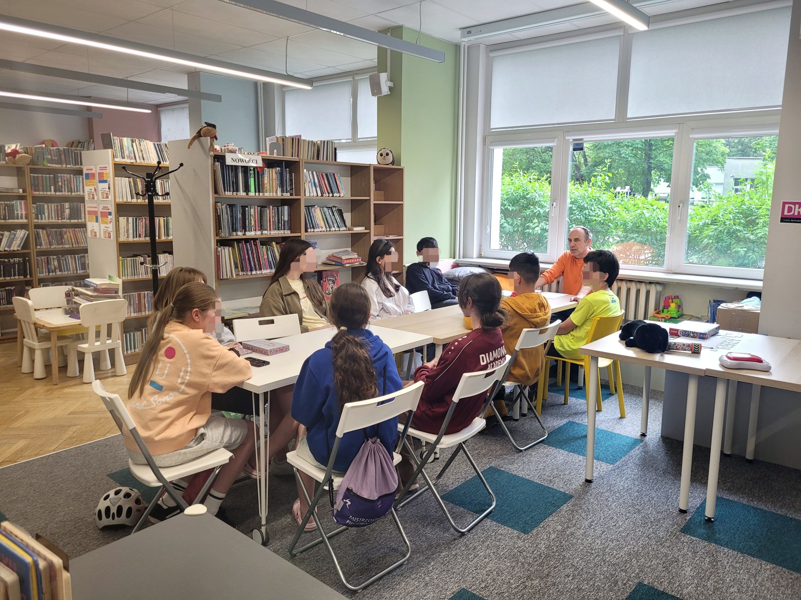 Spotkanie Młodzieżowego Dyskusyjnego Klubu Książki w przestrzeni bibliotecznej. Grupa około ośmiorga nastolatków siedzi wokół złączonych białych stołów w nowoczesnych, jasnych wnętrzach. Po lewej stronie w tle widoczne są wysokie regały pełne książek oraz stolik dziecięcy z małymi krzesełkami. Na końcu stołu, przy dużym oknie wychodzącym na zielone podwórko, siedzi mężczyzna prowadzący spotkanie w pomarańczowej bluzce i rozmawia z młodzieżą