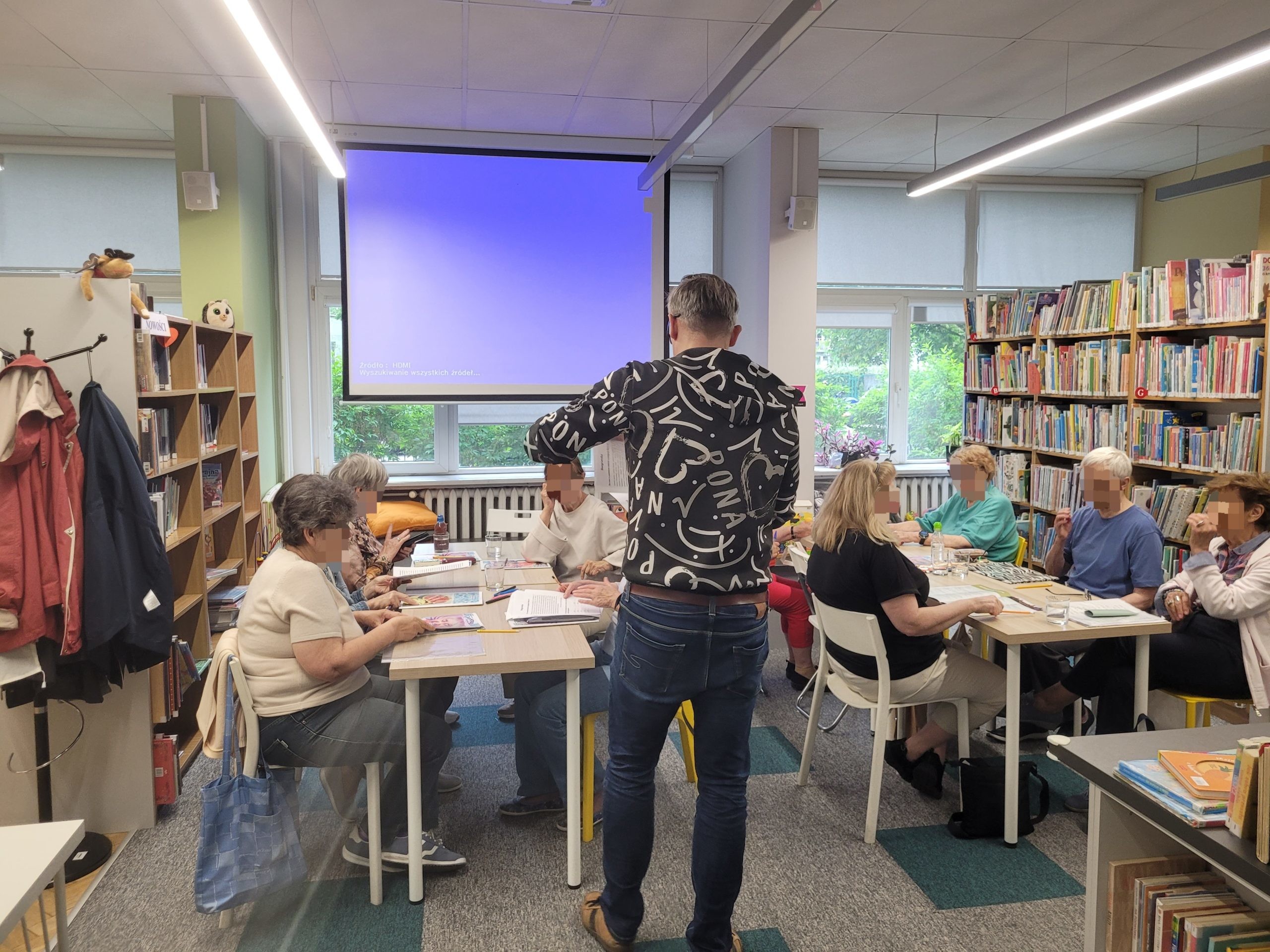 Uczestnicy klubu seniora siedzą przy stolikach w przestrzeni bibliotecznej, zajęci pracą pisemną. Na pierwszym planie, tyłem do obiektywu, stoi prowadzący w ciemnej bluzie z charakterystycznymi białymi napisami. W tle widoczny jest duży, podświetlony ekran projekcyjny oraz wysokie regały gęsto zapełnione kolorowymi książkami dla dzieci i młodzieży.