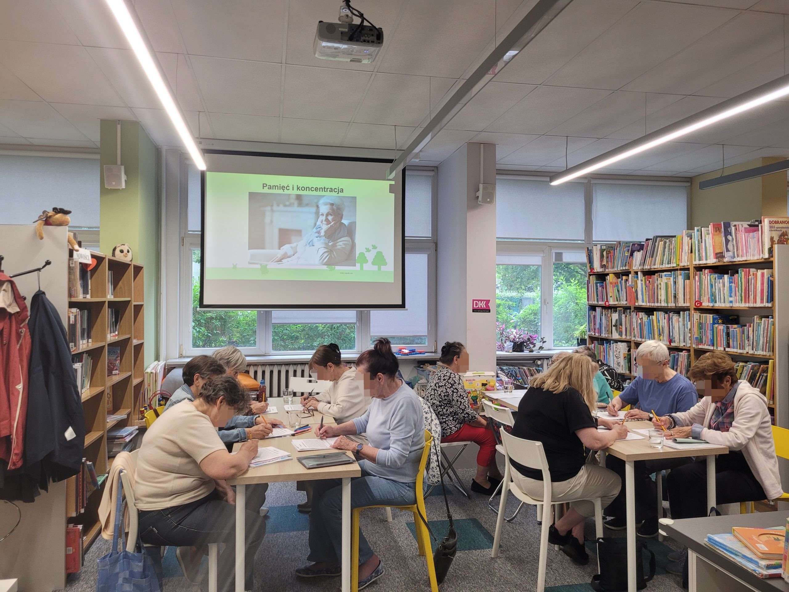 Kolejne ujęcie z warsztatów „Pamięć i koncentracja” w bibliotece. Na ekranie widnieje slajd przedstawiający starszą, uśmiechniętą kobietę podpierającą podbródek. Pod ekranem, przy stołach, grupa seniorek i seniorów w skupieniu wypełnia testy lub zadania logiczne za pomocą ołówków. Wokół znajdują się regały z książkami.