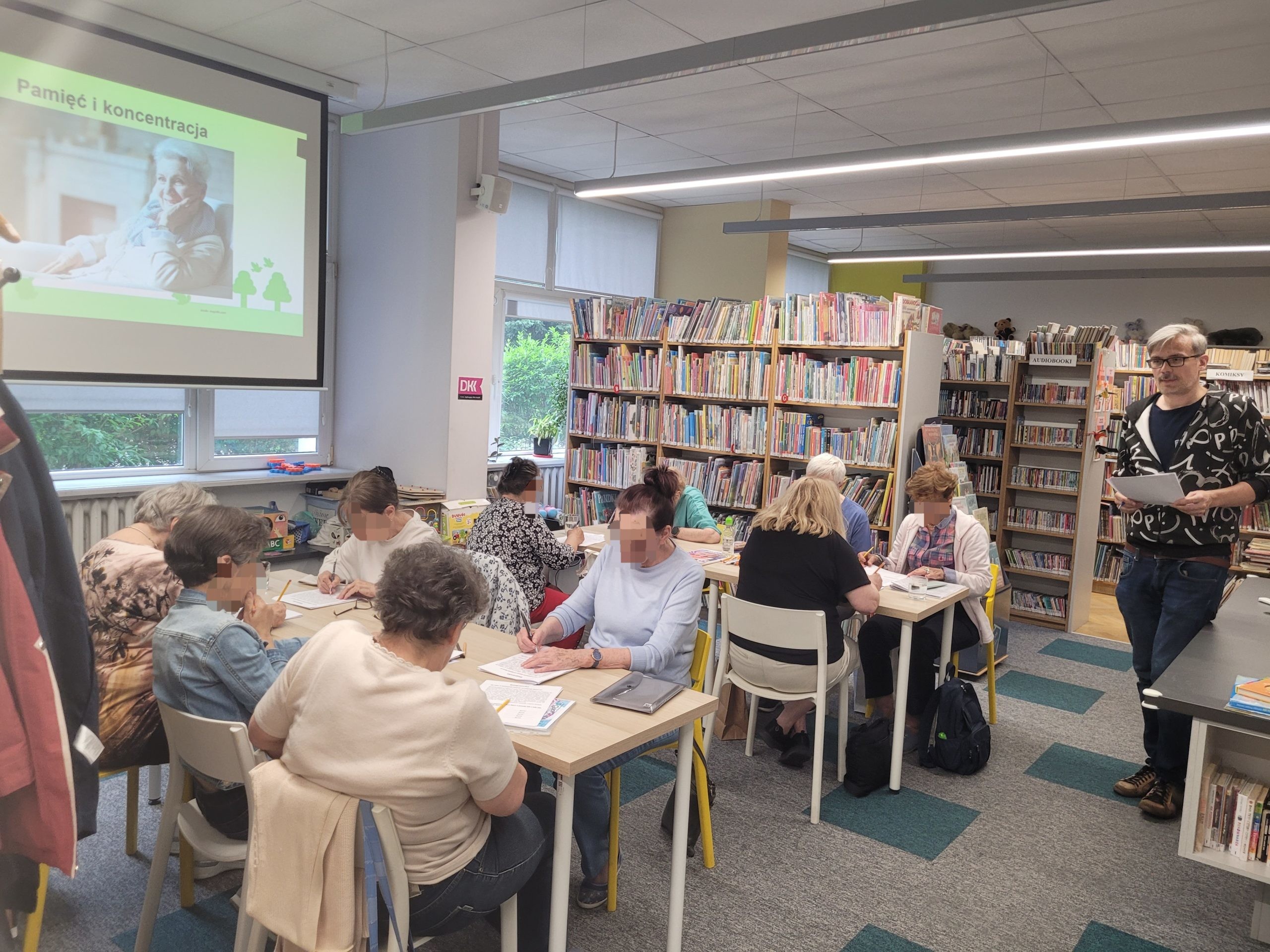 Perspektywa z boku sali bibliotecznej podczas warsztatów dla seniorów. Po lewej stronie stoi prowadzący zajęcia z kartką w ręku. Uczestnicy siedzą przy długich stołach, pochyleni nad arkuszami papieru i piszący ołówkami. W tle wyświetlany jest slajd z prezentacji o pamięci, a przestrzeń otaczają regały wypełnione literaturą.
