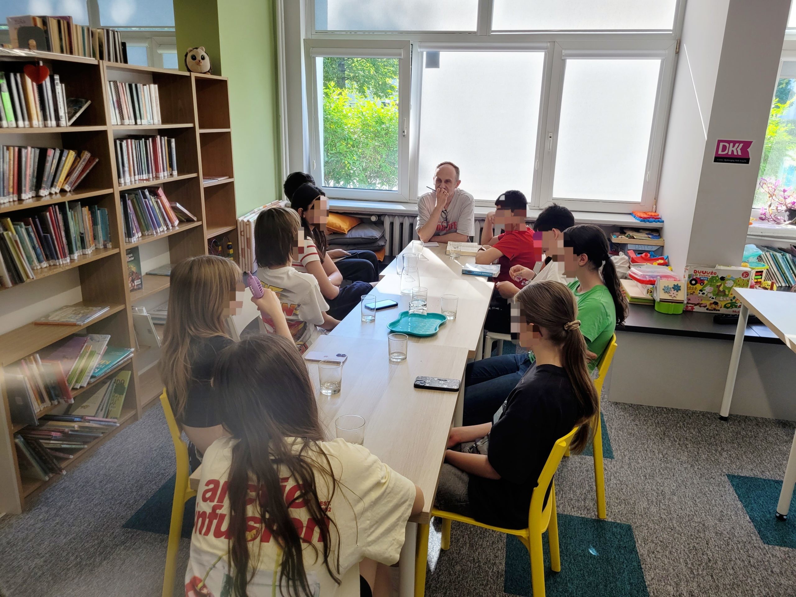 Spotkanie Młodzieżowego Dyskusyjnego Klubu Książki w jasnym pomieszczeniu biblioteki. Grupa młodzieży siedzi przy długim, jasnym stole na żółtych krzesłach. Na końcu stołu, tyłem do okna, siedzi dorosły prowadzący, który rozmawia z uczestnikami. Po lewej stronie widoczne są wysokie, drewniane regały wypełnione książkami, a po prawej kącik dla dzieci z zabawkami oraz logo DKK na filarze. Na stole stoją szklanki z wodą.