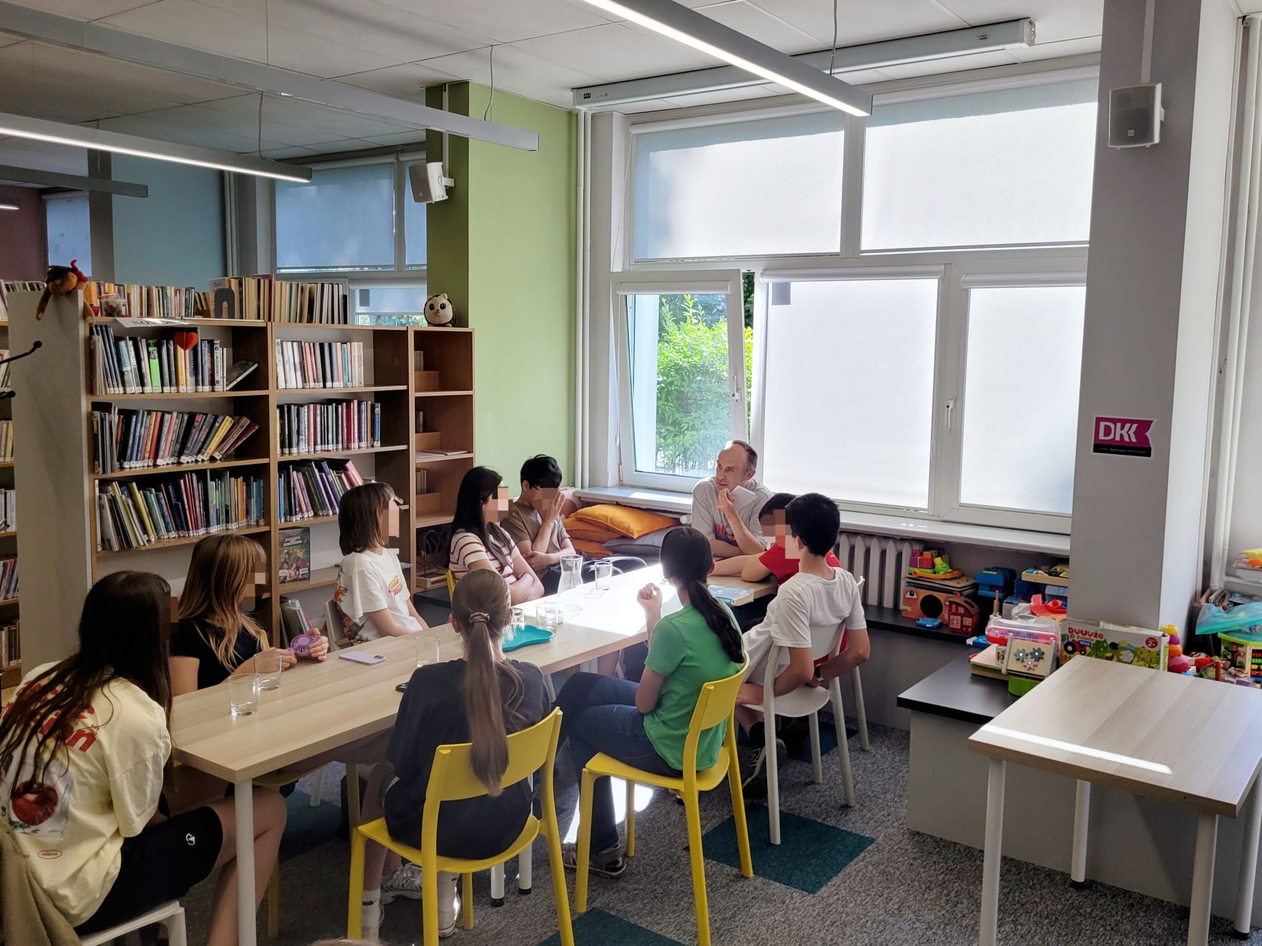 Szeroki kadr przedstawiający spotkanie klubowe w bibliotece. Około dziesięcioro nastolatków siedzi wokół złączonych jasnych stołów, słuchając mężczyzny prowadzącego dyskusję, który gestykuluje ręką. Wnętrze jest nowocześnie oświetlone podłużnymi lampami sufitowymi. Po lewej stronie stoją regały z książkami, a duże okna w tle wpuszczają naturalne światło. Przy ścianie po prawej widoczny jest regał z kolorowymi zabawkami.