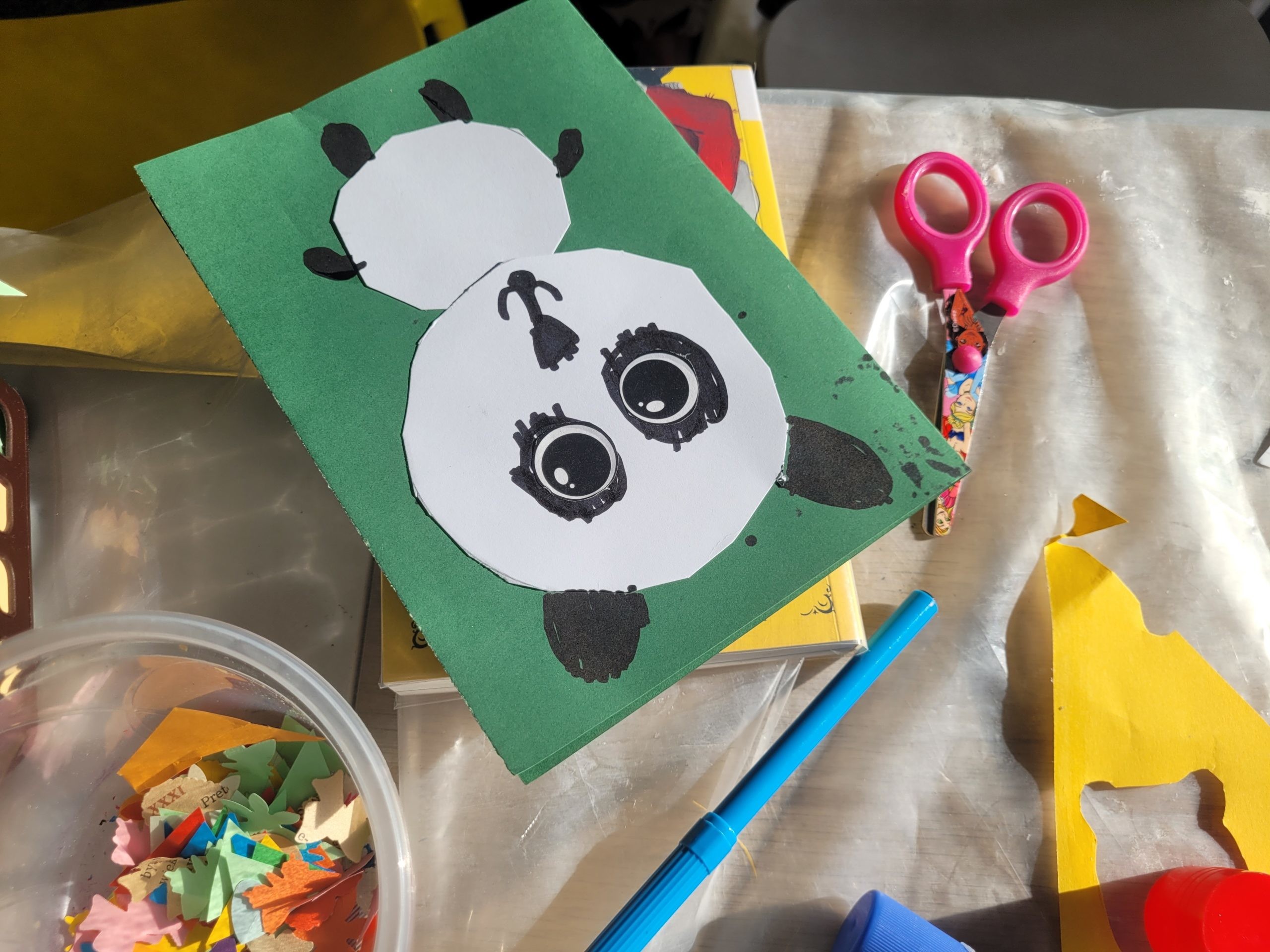 Widok z góry na zieloną laurkę z motywem pandy leżącą na stole obok różowych nożyczek i niebieskiego pisaka. Panda została wykonana z naklejonych na siebie białych kół z papieru z dorysowanymi czarnym markerem uszami, łapkami oraz dużymi oczami. W prawym górnym rogu kadru widoczna jest miseczka z drobnymi, kolorowymi ścinkami papieru.