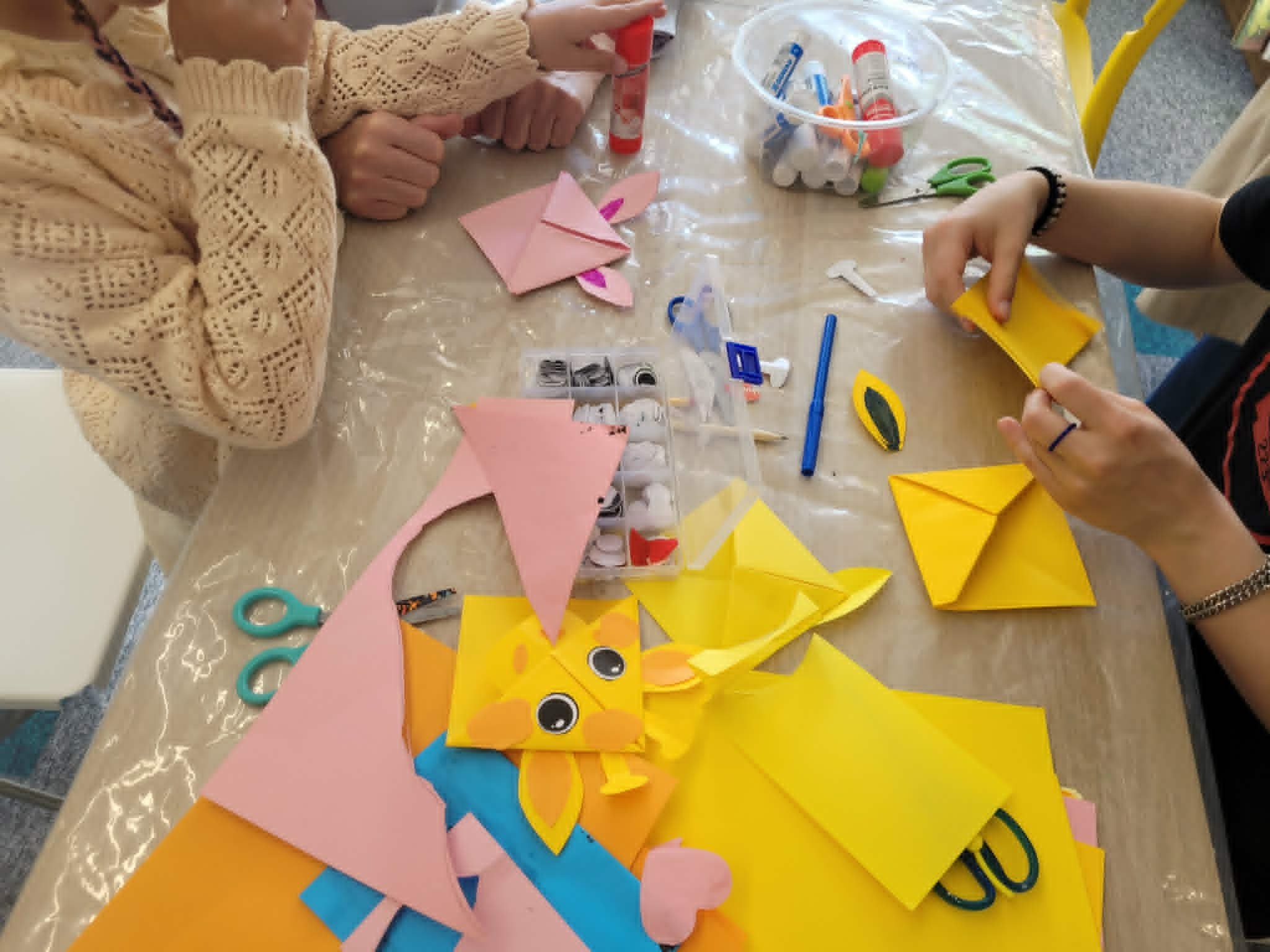Widok z góry na stół pokryty folią, przy którym dzieci składają papierowe zakładki origami. Na środku stołu leży gotowa żółta zakładka w kształcie głowy żyrafy z ruchomymi oczkami, a obok niej różowa zakładka z uszami królika. Wokół rozsypane są ścinki żółtego i różowego kartonu, kleje w sztyfcie, nożyczki oraz plastikowy organizer z plastikowymi oczkami do naklejania.