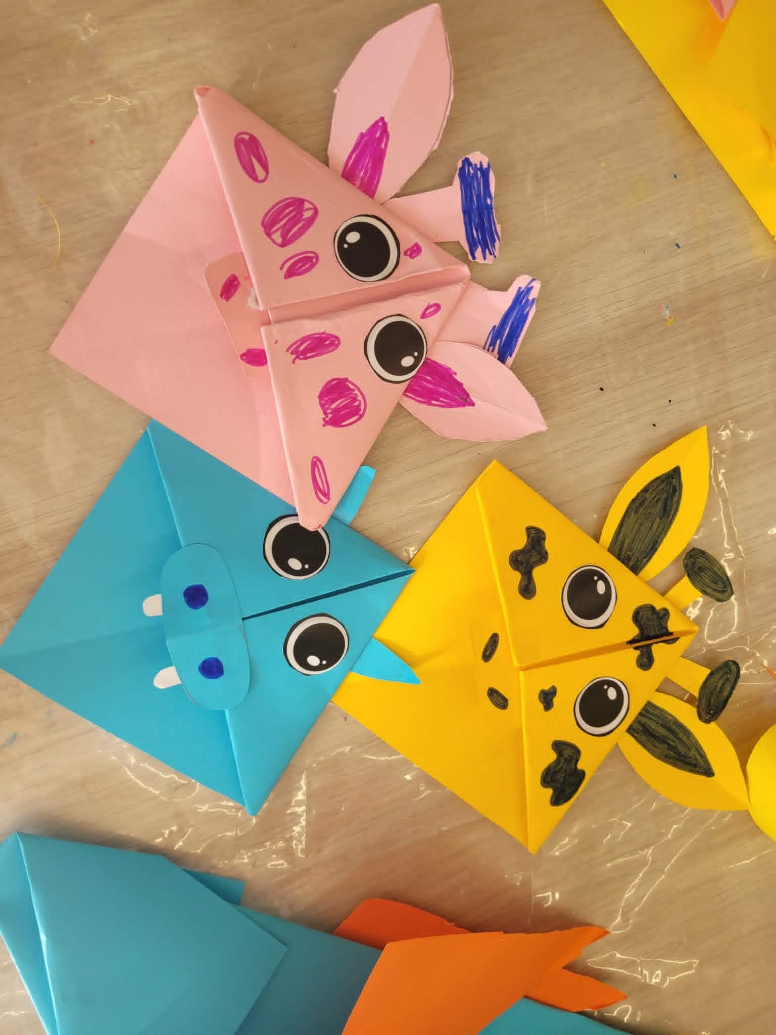 Trzy kwadratowe zakładki narożne do książek wykonane techniką origami, leżące obok siebie na jasnym blacie. Górna, różowa zakładka przypomina świnkę lub potworka z fioletowymi plamami. Lewa, niebieska przedstawia hipopotama z białymi zębami, a prawa, żółta to głowa żyrafy z domalowanymi czarnymi łatkami, rogami i uszami.