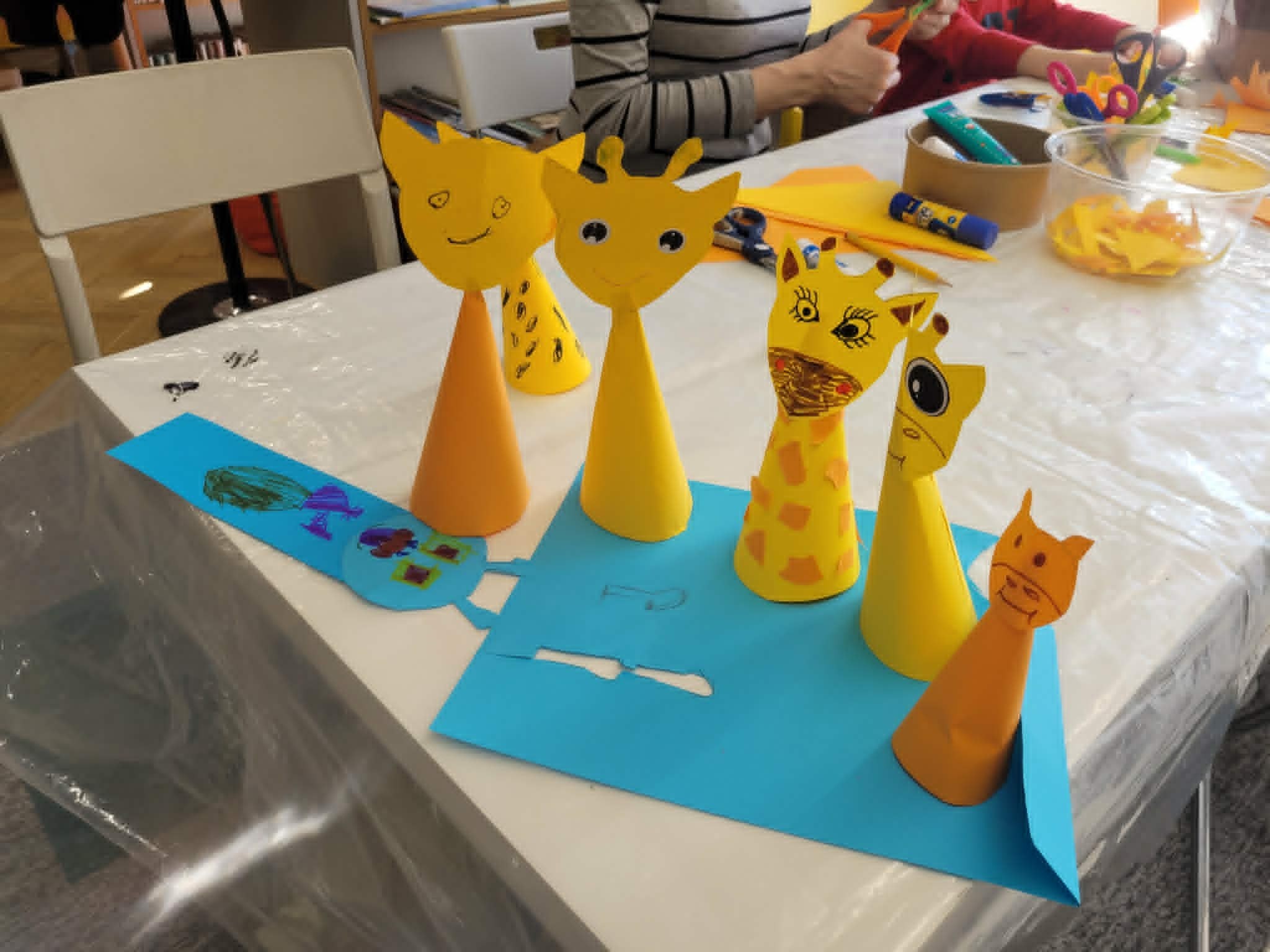 Rząd sześciu gotowych, przestrzennych figurek zwierząt wykonanych z papierowych stożków, ustawionych na niebieskiej podkładce. Wśród prac znajdują się cztery żółto-pomarańczowe żyrafy o różnych wzorach łat i wyrazach pyszczków narysowanych mazakami oraz jedna figurka przypominająca lisa. W tle widoczna jest pracująca przy stole dorosła osoba oraz regały z książkami.