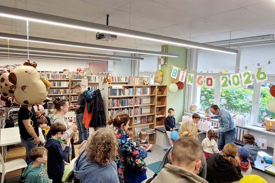Ogólne ujęcie gali Bingo 2026. Bibliotekarz rozmawia z dziećmi przy stoliku z nagrodami. Na środku chłopiec bawi się niebieskim balonem, a inne dzieci czekają na swoją kolej. W tle kolorowy napis "BINGO 2026".