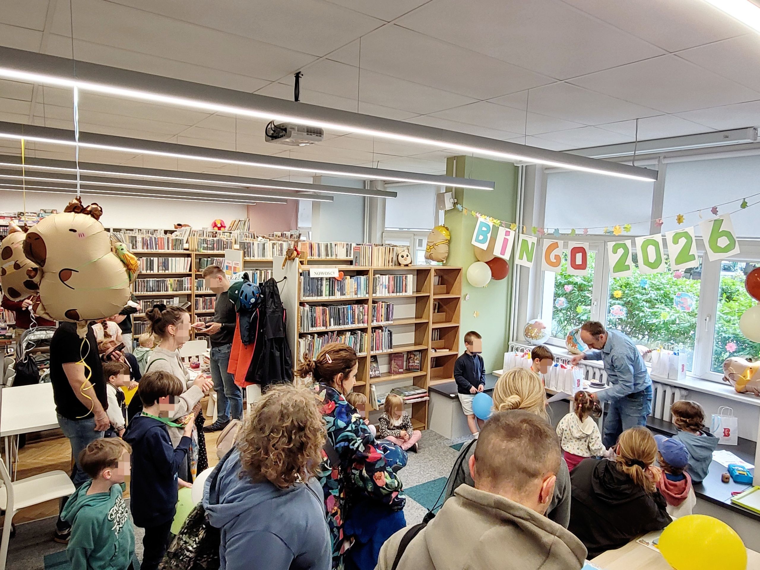 Ogólne ujęcie gali Bingo 2026. Bibliotekarz rozmawia z dziećmi przy stoliku z nagrodami. Na środku chłopiec bawi się niebieskim balonem, a inne dzieci czekają na swoją kolej. W tle kolorowy napis "BINGO 2026".