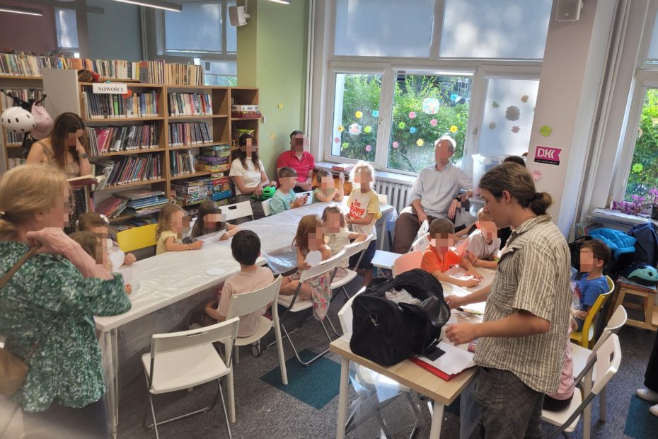 Szersze ujęcie sali bibliotecznej wypełnionej dziećmi i dorosłymi podczas kreatywnych warsztatów. Po lewej stronie, wzdłuż ściany z wysokimi regałami pełnymi książek, kilkoro dzieci siedzi przy stole pokrytym folią ochronną, zajętych pracą plastyczną. W tle, przy oknie udekorowanym kolorowymi kwiatami, widać dorosłych opiekunów obserwujących zajęcia. Po prawej stronie zdjęcia młody instruktor stoi przy oddzielnym stoliku, przy którym siedzi kolejna grupa dzieci, i porządkuje materiały w otwartej czarnej torbie. Całość oddaje atmosferę zaangażowania i twórczej zabawy w przyjaznej, oświetlonej naturalnym światłem przestrzeni biblioteki.