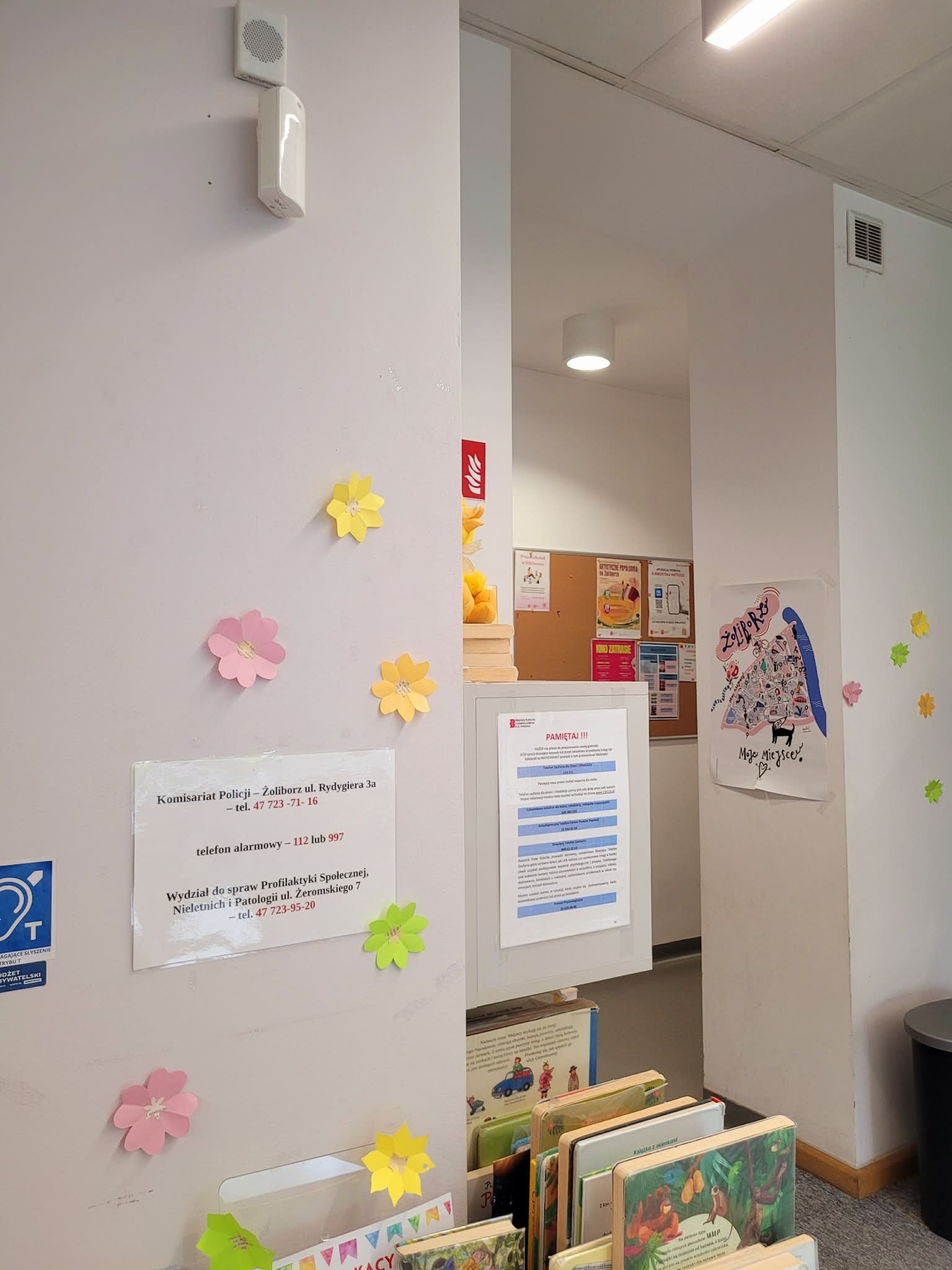 Na ścianie biblioteki przy wejściu zamontowany jest znacznik systemu TOTUPOINT. W tle widoczne są regały z książkami dla dzieci, tablica informacyjna oraz plakaty. System ułatwia orientację w przestrzeni osobom z niepełnosprawnością wzroku.