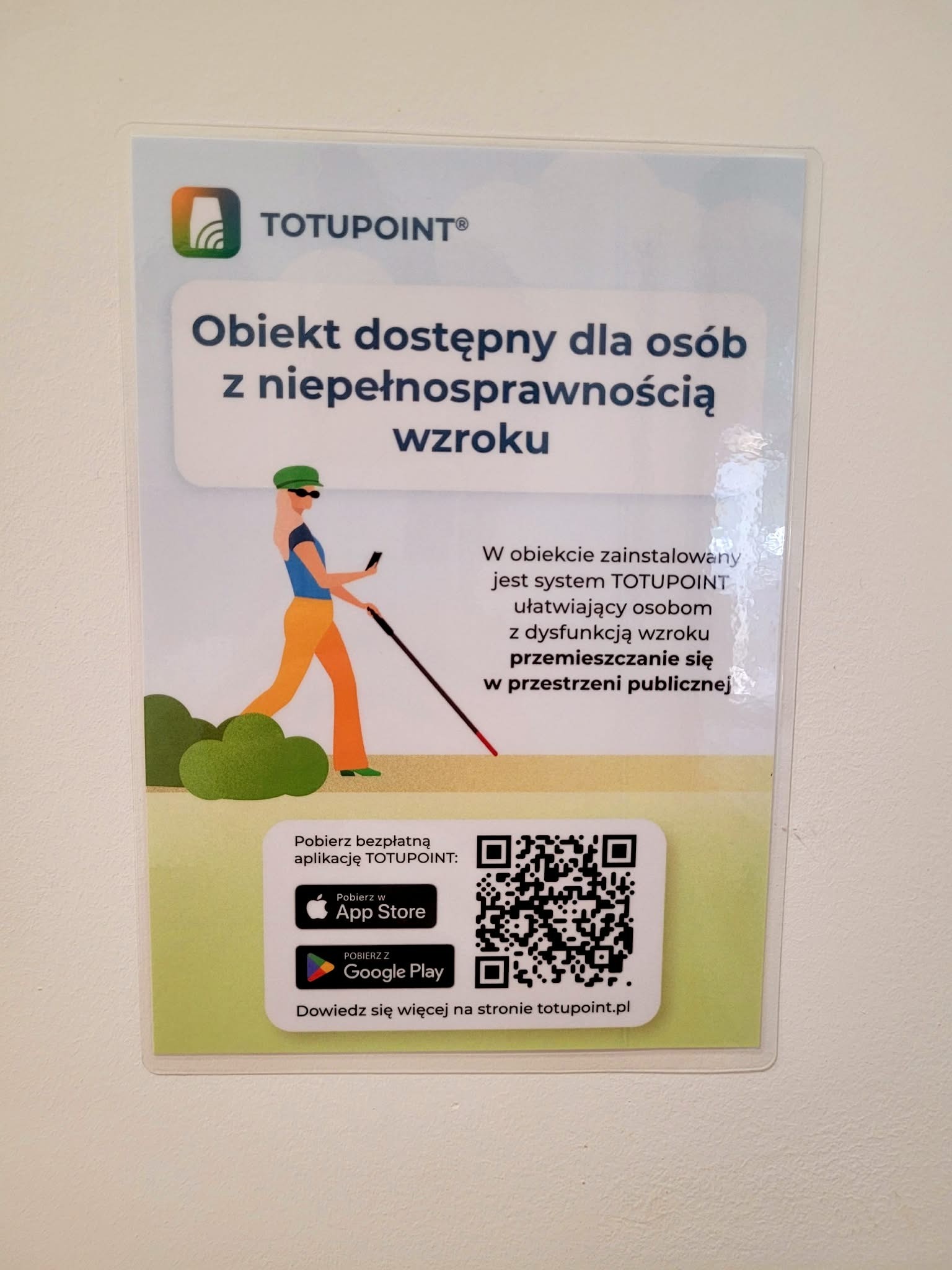 Na jasnej ścianie biblioteki znajduje się plakat informujący o dostępności obiektu dla osób z niepełnosprawnością wzroku. W górnej części umieszczono logo systemu TOTUPOINT oraz napis „Obiekt dostępny dla osób z niepełnosprawnością wzroku”. Poniżej znajduje się ilustracja osoby poruszającej się z białą laską i smartfonem oraz informacja, że w obiekcie zainstalowano system TOTUPOINT ułatwiający osobom z dysfunkcją wzroku samodzielne poruszanie się w przestrzeni publicznej. W dolnej części plakatu widoczne są kody QR oraz przyciski do pobrania bezpłatnej aplikacji TOTUPOINT z App Store i Google Play, a także adres strony internetowej systemu.