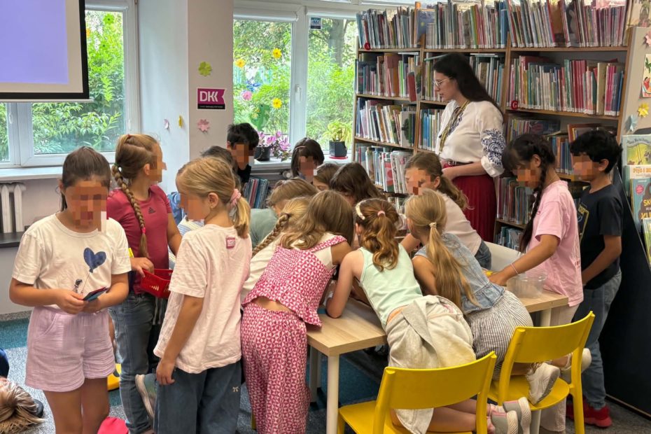 Grupa dzieci stoi wokół stołu w bibliotece, pochylając się nad materiałami plastycznymi. Obok stołu stoi prowadząca zajęcia kobieta w białej bluzce z haftem, a w tle widać regały pełne książek.